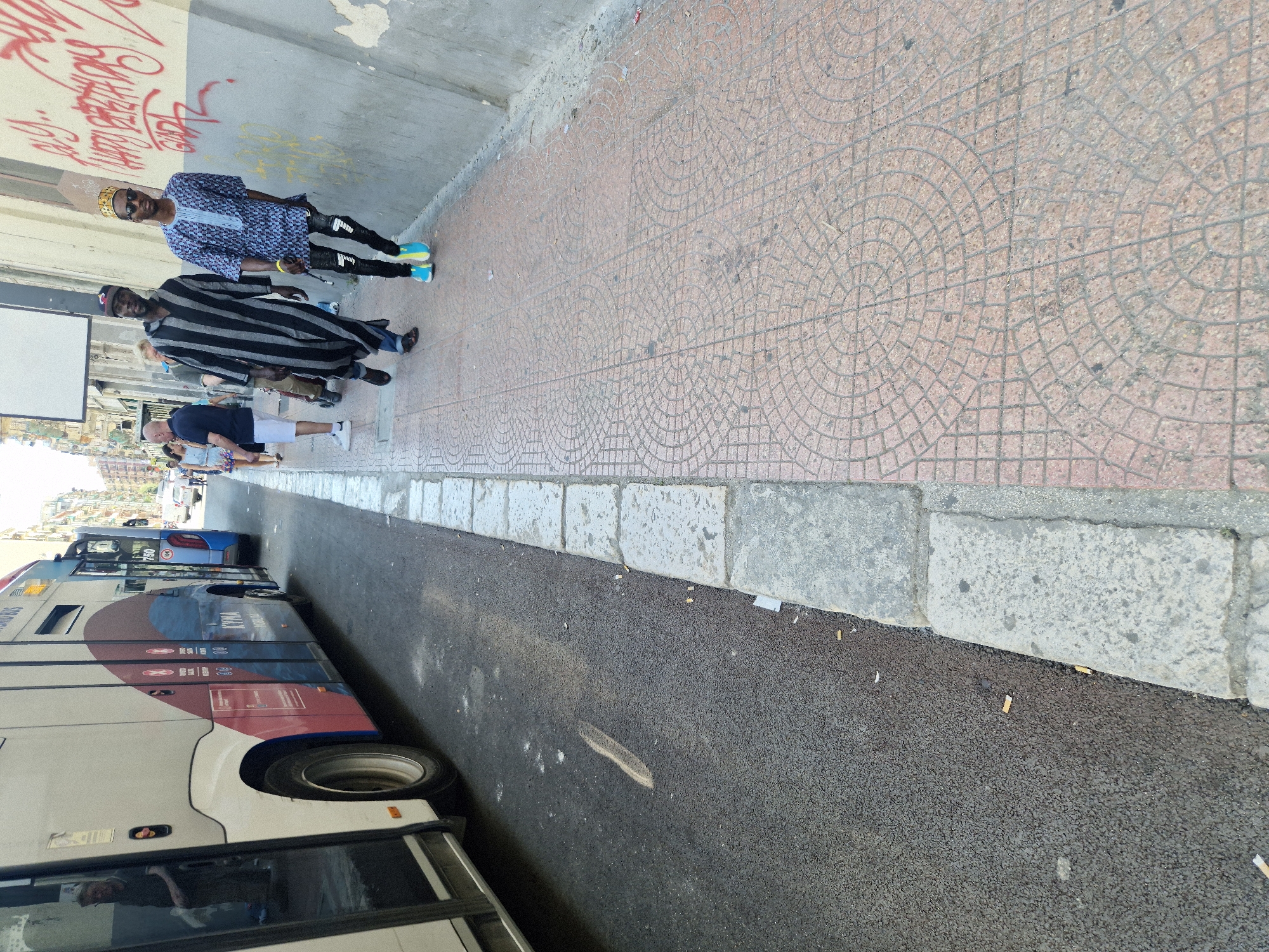 This image depicts a street scene in Taranto, Italy. In the foreground, a bus occupies the left side of the frame. To the right, a sidewalk is visible, marked by a distinctive reddish-brown tile pattern. Two men are walking along the sidewalk. The man on the left wears a long, black and white striped garment and a dark baseball cap. The man on the right is dressed in a blue patterned robe and a gold crown-like headpiece, paired with dark pants and bright blue and yellow sneakers. The background shows buildings lining the street. The overall impression is of a sunny day in an urban environment.