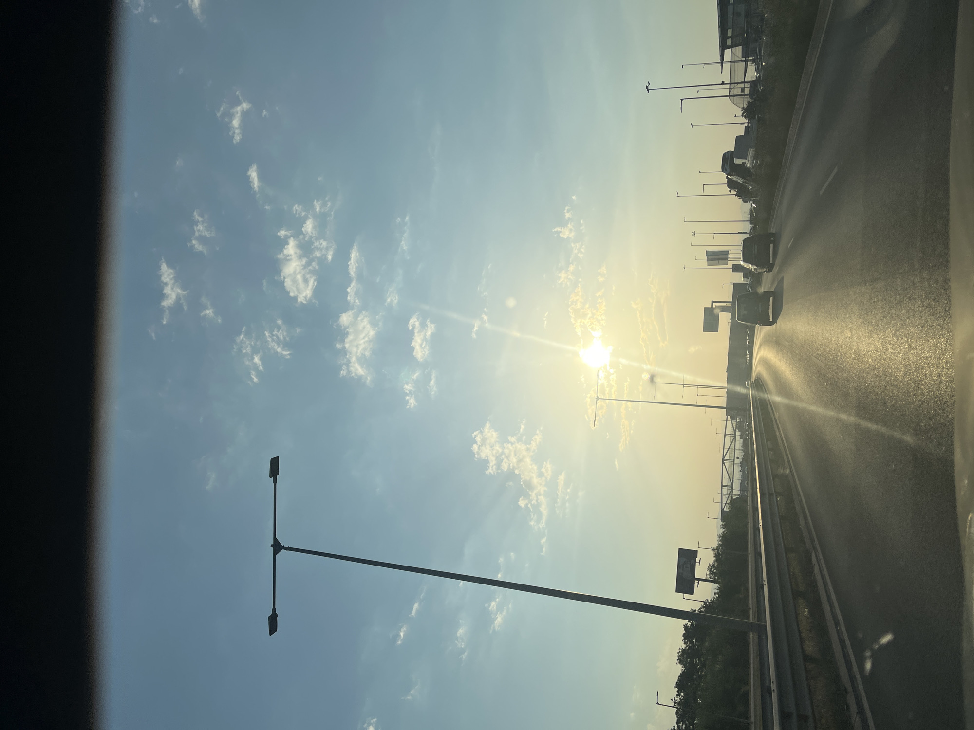 A view from a moving vehicle on a highway during sunset. The sky is a gradient of blue and yellow, with scattered white clouds. Sunlight beams through the clouds, creating lens flare. On the left side of the highway, there is a tall street lamp and trees. On the right side, there are more street lamps, some parked cars, and buildings in the distance. Several cars are visible on the highway ahead. The road surface is asphalt with white lane markings. The overall scene suggests travel and a journey towards the setting sun.