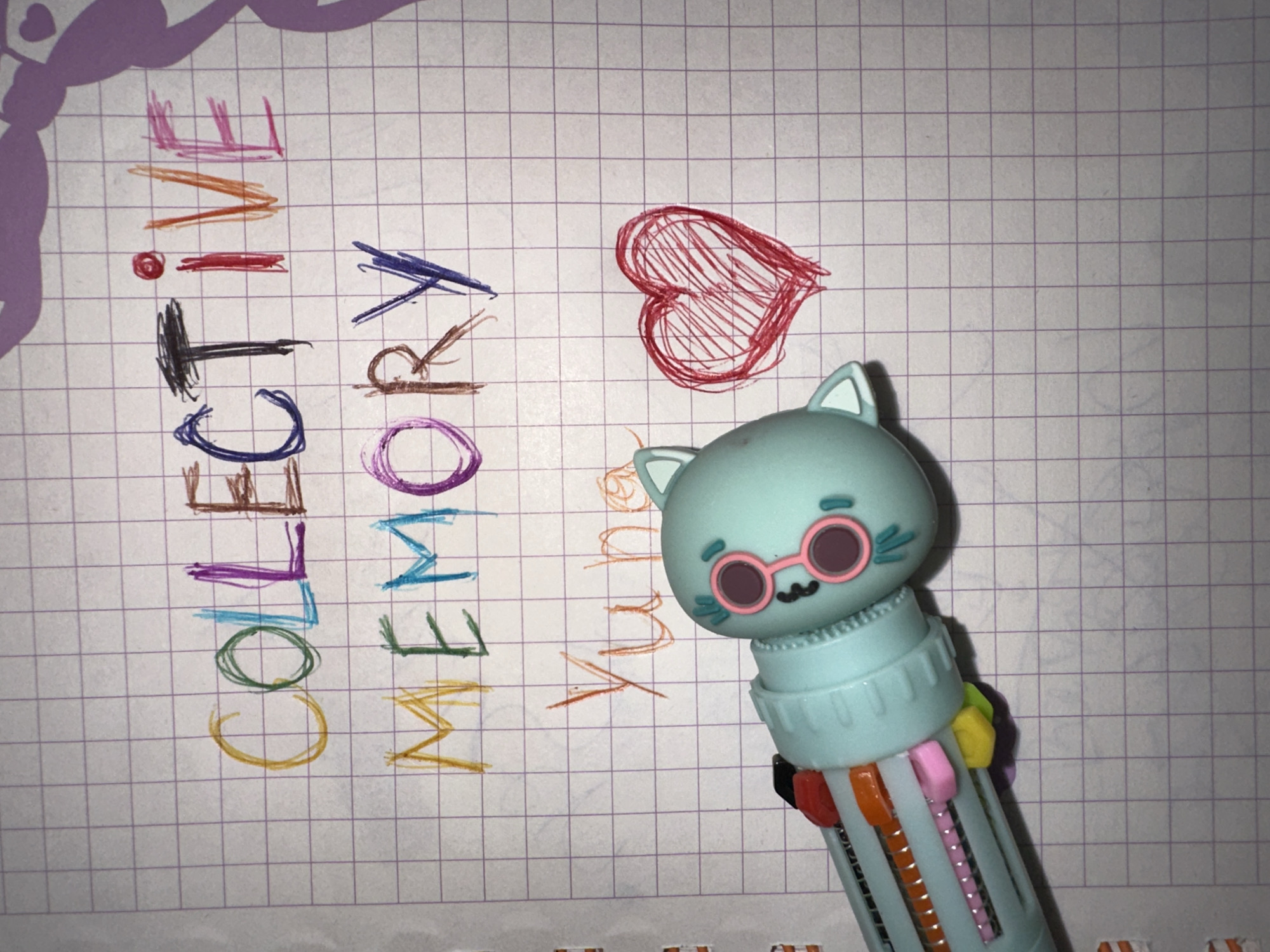 A sheet of grid paper rests on a surface, displaying the words "COLLECTIVE" and "MEMORY" written in different colored markers. A red heart drawing is positioned above the word "MEMORY". A blue multi-color pen, with a cat-shaped topper wearing glasses, is present on the right side of the paper. The setting appears to be indoors. There is no indication that the image was taken in Esenyurt, Turkey.
