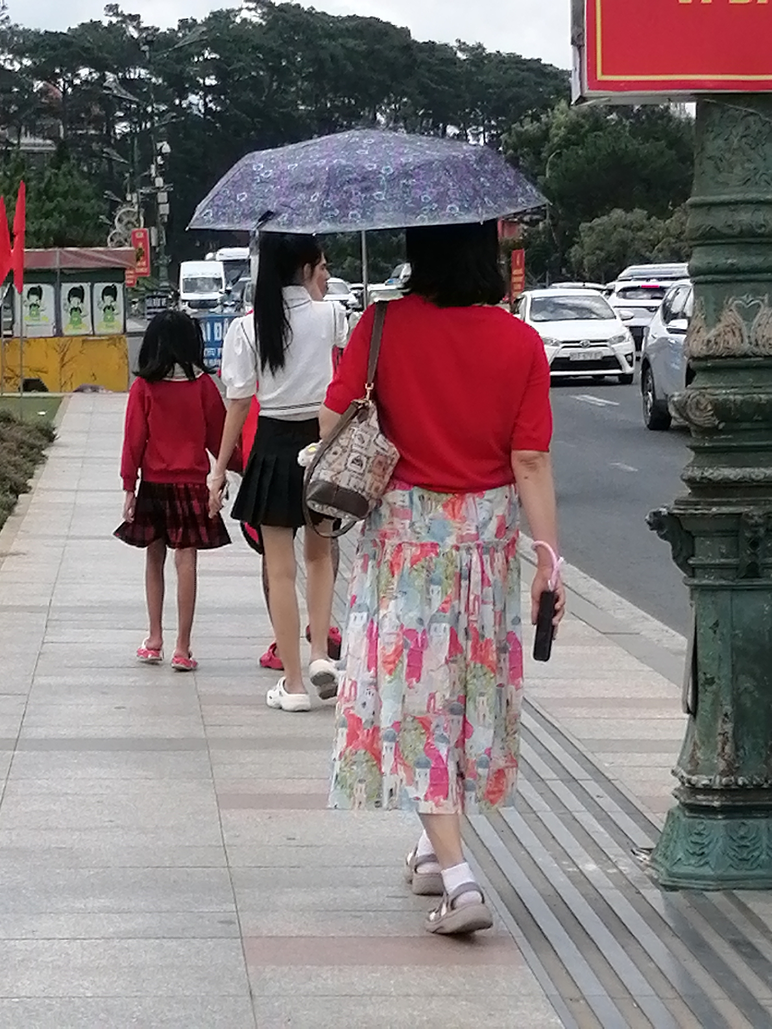 The image shows a group of people walking on a sidewalk in Da Lat, Vietnam. A woman in a red shirt and a floral skirt holds an umbrella over herself and a younger girl in a red sweater and plaid skirt. The girl is walking in front of the woman, holding her hand. Another woman in a white shirt and black skirt walks beside the girl, her hand on the girl's shoulder. The sidewalk is paved with light gray tiles. In the background, trees line the street, and cars are visible on the road. The overall scene appears to be a street in a city on a cloudy day.