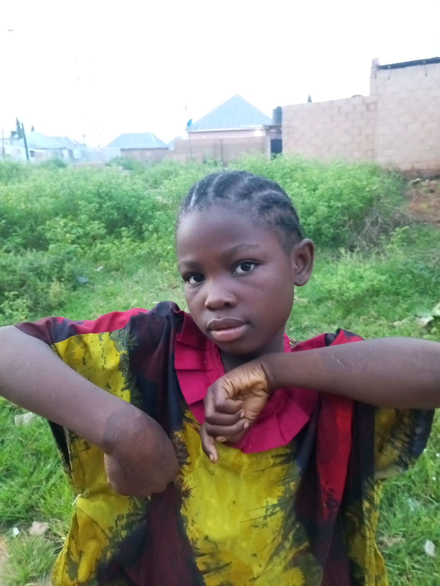 A young girl with braided hair, wearing a yellow and red patterned garment, stands outdoors with a slightly turned head and a neutral expression. Her right arm is bent across her chest, with her hand resting on her left arm. She appears to be in an open area with green vegetation in the background, and buildings are visible in the distance. The overall setting suggests a rural or semi-urban environment in Tempure, Nigeria.