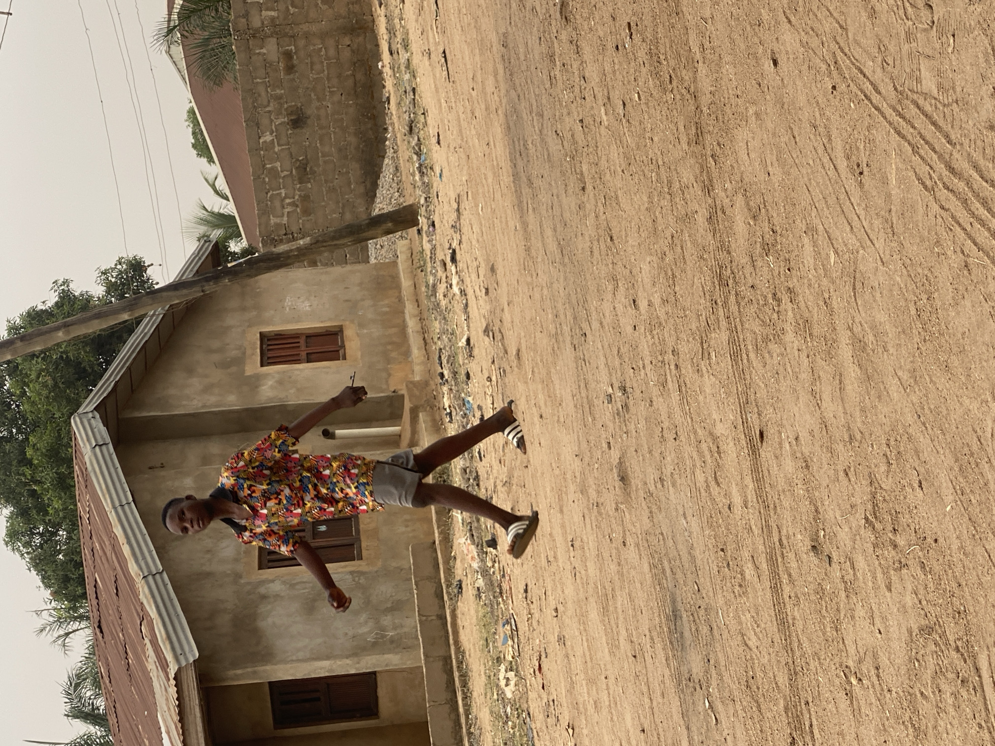 A young individual stands on a sandy, unpaved road in Sabon Tasha, Nigeria. The person is wearing a colorful, patterned shirt and gray shorts, with sandals on their feet. Behind them is a building with a light-colored facade and windows. The environment includes trees, a partially visible rooftop, and an adjacent brick structure. The dirt road shows tire tracks and some scattered debris.