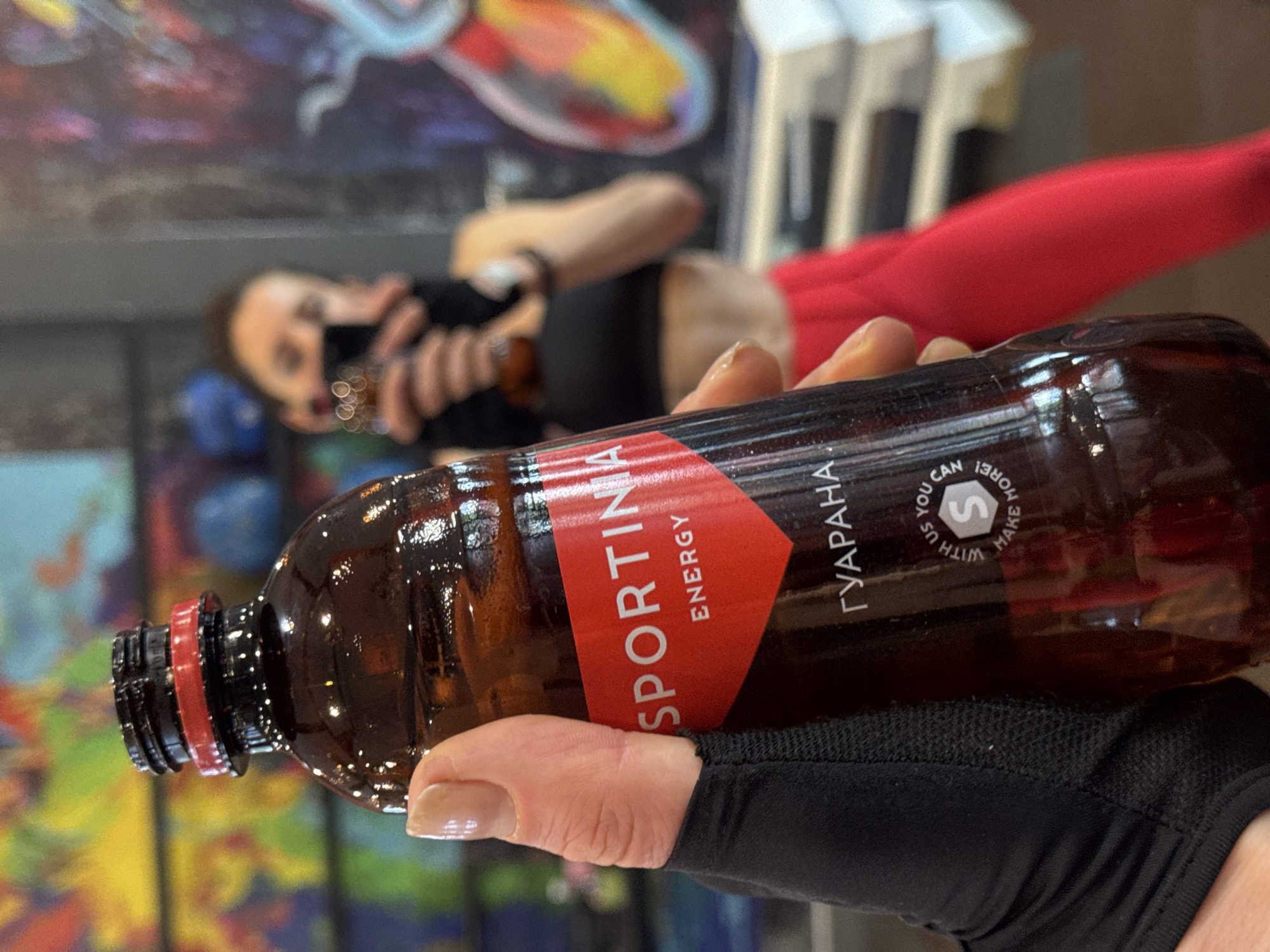 A person, wearing a black top and red pants, is holding an energy drink. The label on the bottle indicates "SPORTINA ENERGY" with "ГУАРАНА" (Guarana) written in Cyrillic script. The background includes an abstract painting and gym equipment. The location is potentially Vladikavkaz, Russia, based on the Cyrillic script on the bottle.