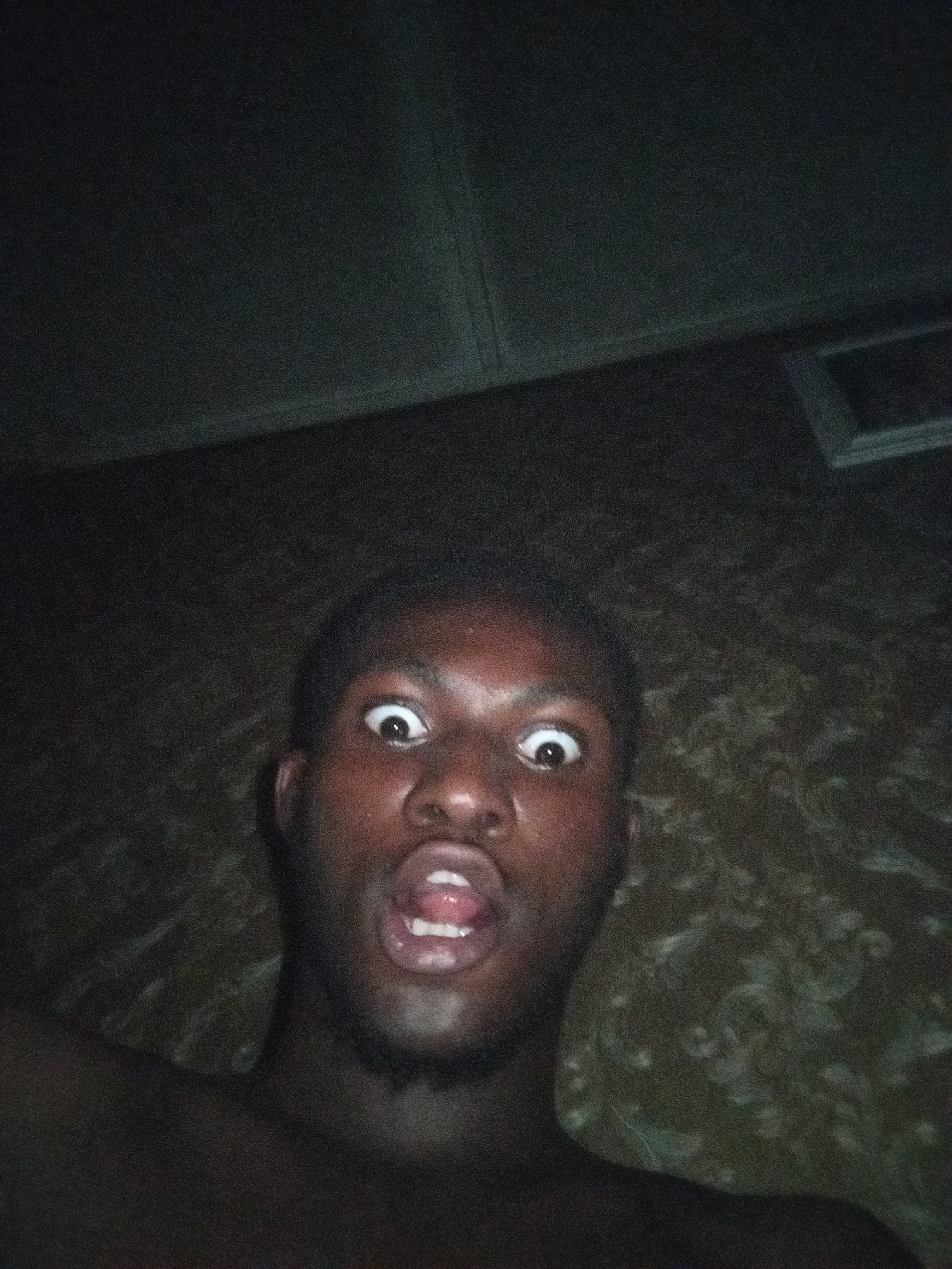 A low-angle selfie captures a Black male indoors. The man is shirtless with dark skin, and his eyes are wide open. His mouth is open, revealing his teeth and tongue. Behind him, the background is dark with a patterned wall and a ceiling with a rectangular object. Location: Unknown, Unknown.