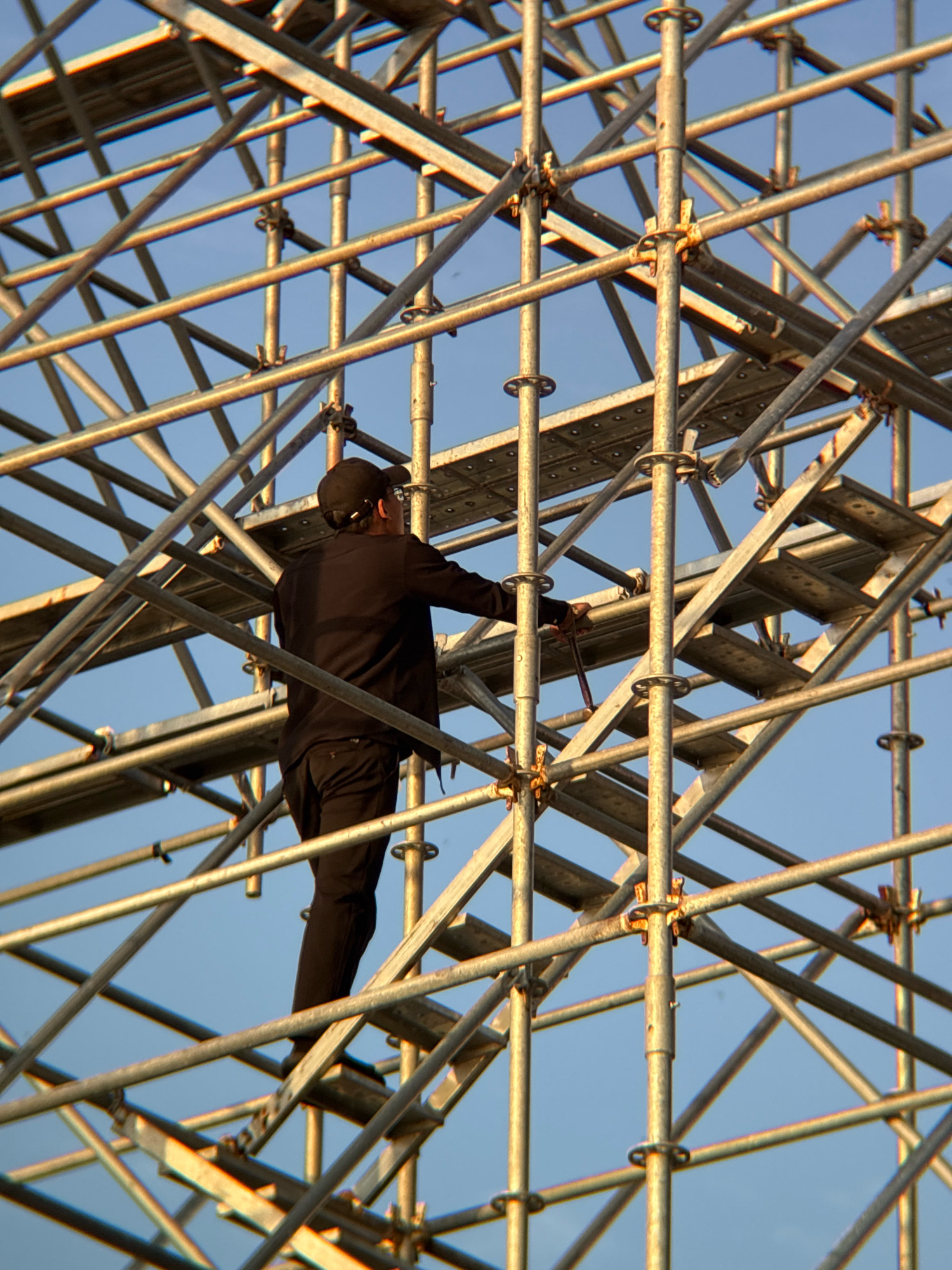 A worker in a black shirt and pants, wearing a black baseball cap, is ascending a scaffolding structure. They are holding a tool, possibly a wrench, in their right hand as they navigate the metal framework against a clear blue sky. The scaffolding is extensive and appears to be part of a construction or renovation project. The image is taken from a low angle, looking up at the worker and the intricate metal grid of the scaffolding. The scene is devoid of any specific landmarks to definitively place it in Nha Trang, Vietnam, however, it is a common sight in urban development areas globally.