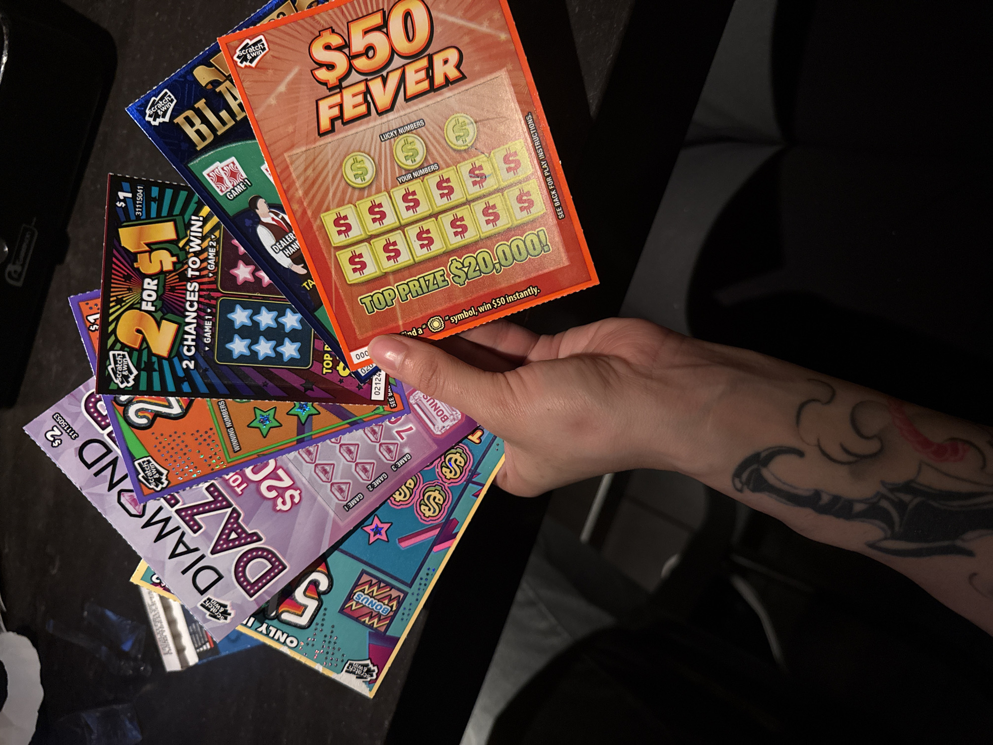 A person is holding a stack of lottery scratch-off tickets. The tickets are various colors and designs, including "Diamond Daze", "2 for $1", "Blackjack", and "$50 Fever." The "$50 Fever" ticket advertises a top prize of $20,000. The person holding the tickets has a tattoo on their forearm. The tickets are held over a dark surface.
The scene is not identifiable as being in District of North Vancouver, Canada, based on visual elements.