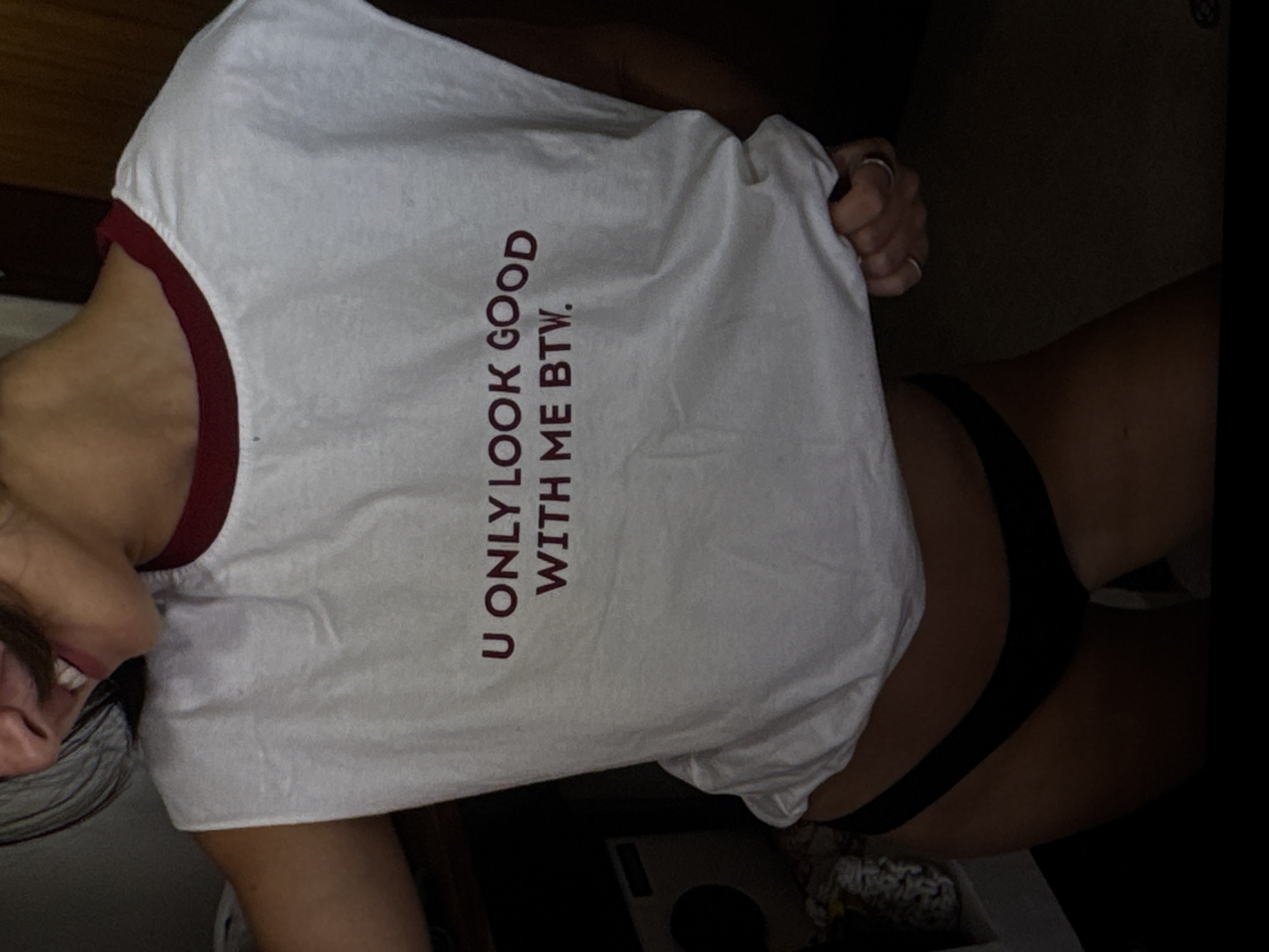 A person is seen from the chest down, wearing a white sleeveless t-shirt with a maroon trim and the text "U ONLY LOOK GOOD WITH ME BTW." in maroon lettering. They are also wearing black underwear. The background is dark and out of focus, with some objects visible on the left, possibly in a room. The person's hands are clasped in front of them, and they are wearing two rings. The scene appears to be indoors, likely in a private setting. There is no indication of the city of Kuta Selatan, Indonesia.