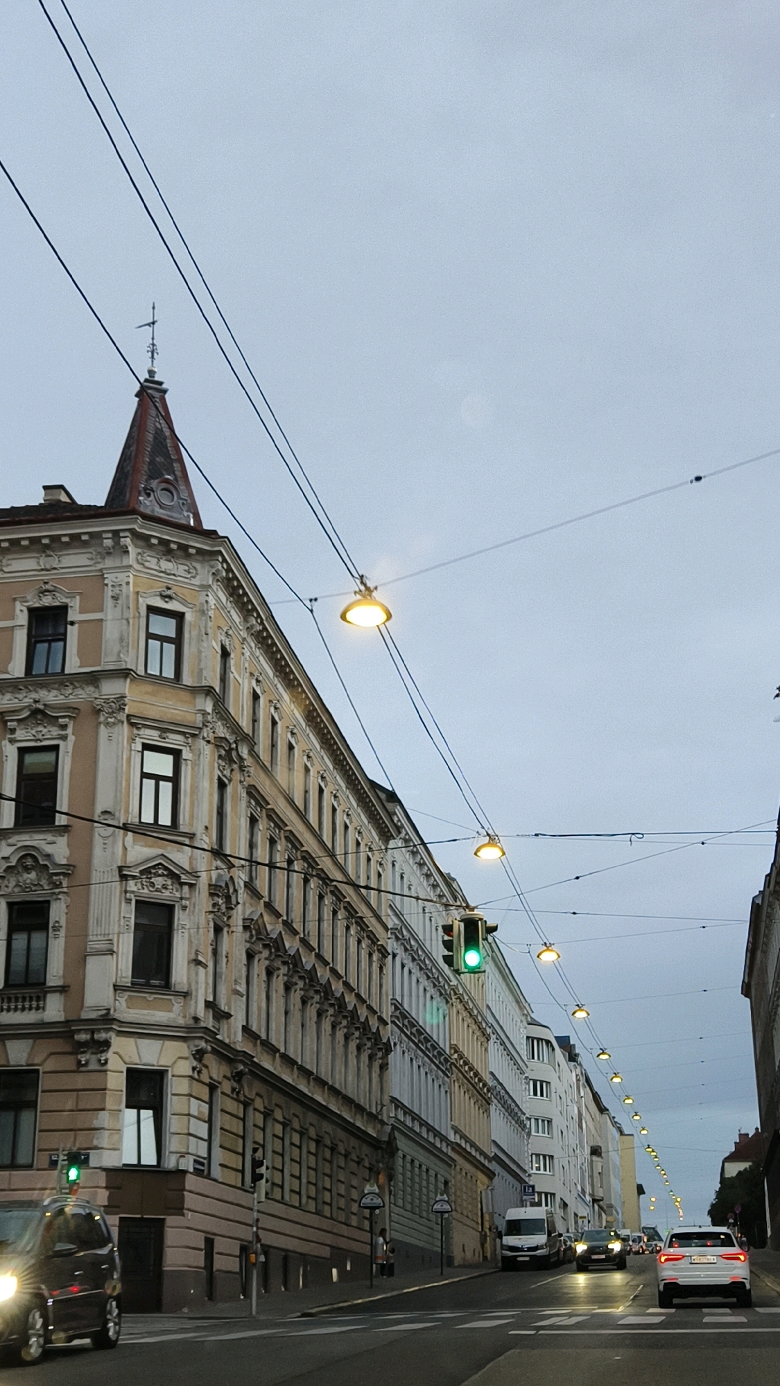 The image depicts a street scene in Vienna, Austria, characterized by an overcast sky and a row of ornate, multi-story buildings. The architecture suggests a European city, with classical detailing on the building facades, including decorative moldings and a prominent tower on the corner building. Overhead, a network of wires stretches across the street, supporting streetlights and traffic signals. One traffic signal is clearly visible and illuminated green. Several vehicles are present on the road, including a dark-colored car in the foreground, a white van, and other cars further down the street. The street itself is paved and marked with pedestrian crossings. The overall impression is of an urban environment during daylight hours, possibly early morning or late afternoon due to the muted lighting conditions.