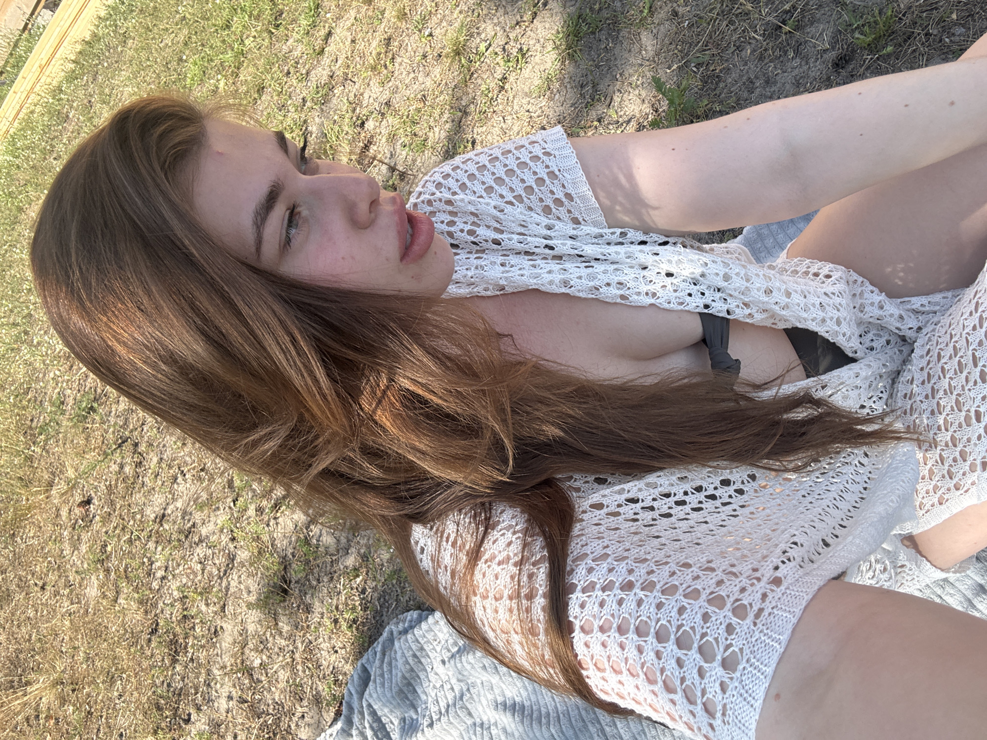 A young woman with long, brown hair lies on her side outdoors. She is wearing a white, crocheted cover-up over a black bra and grey shorts. Her face is partially obscured by her hair, but her eyes are open, and her lips are slightly parted. She appears to be looking up and to the right, with a relaxed expression.
The scene is a grassy area with dry patches and scattered leaves. The sunlight suggests it is daytime, likely a warm day given the woman's attire and the outdoor setting. There are no visible text elements in the image. The presence of what appears to be a wooden structure in the upper left corner might indicate a backyard or a park setting. The overall impression is one of casual relaxation in a natural environment.