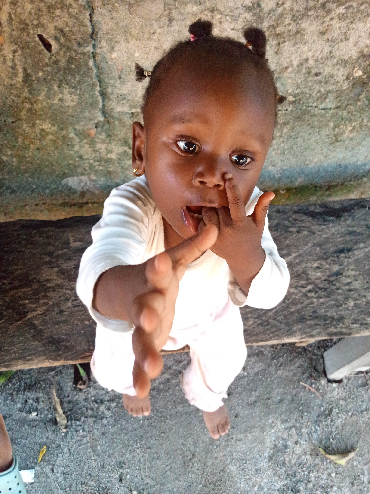 A toddler of African descent, seen from an elevated perspective, stands outdoors in Unknown, Unknown. The child is wearing a light-colored, long-sleeved outfit. The child's hair is styled in small buns. The child's hand is partially inside their mouth and they are extending the other hand towards the camera. The background features a concrete wall.