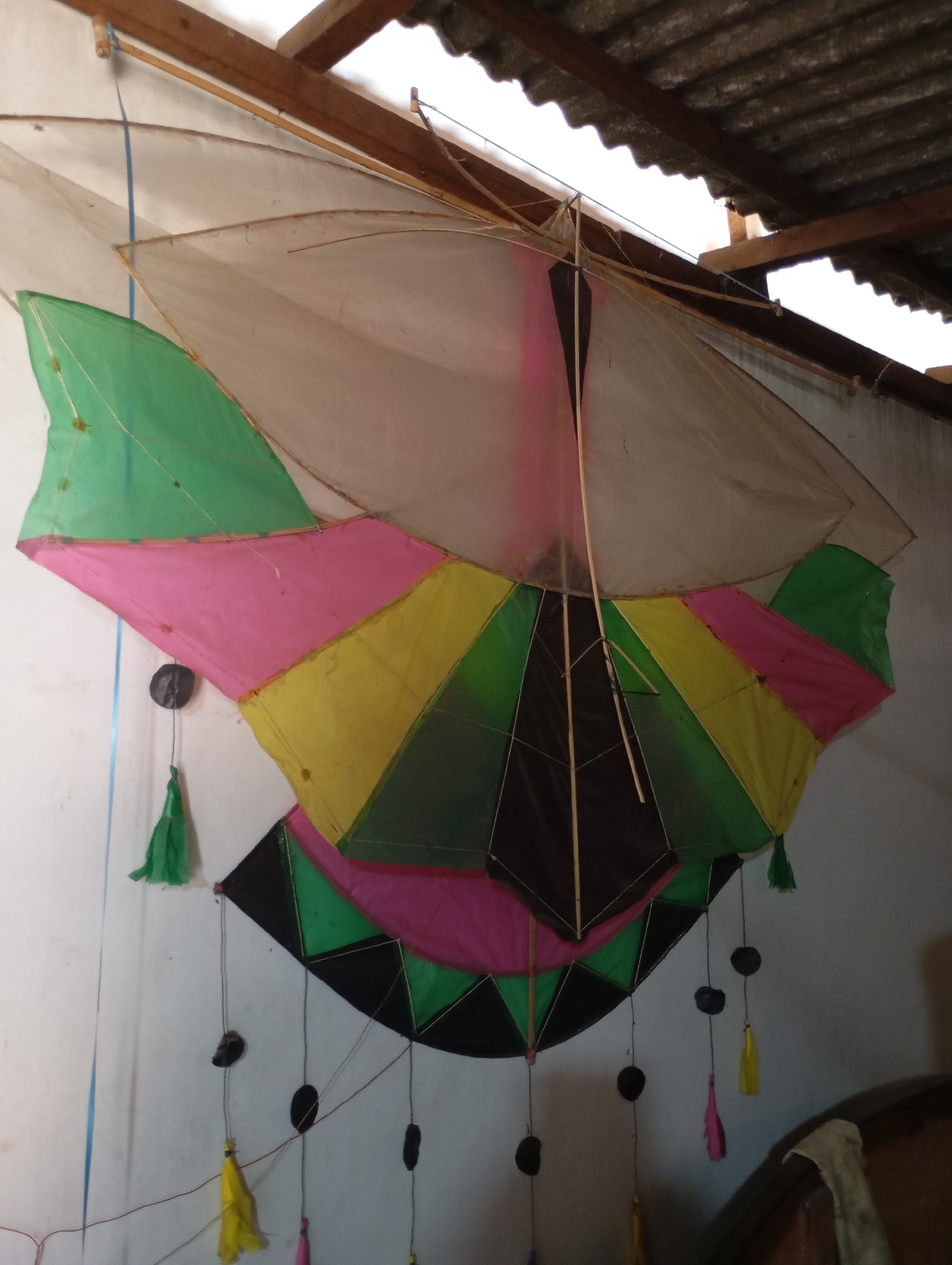 A large, colorful kite is hanging on a white wall, leaning against a wooden beam under a corrugated metal roof. The kite is made of multiple sections of fabric in shades of pink, yellow, green, and black, with a translucent white fabric at the top. Several strings hang from the kite, some with small black weights and colorful tassels in yellow and pink. The environment appears to be outdoors or in a covered outdoor space, with rough textures visible on the wall and roof. The lighting is dim, suggesting it might be late afternoon or a cloudy day. There are no people visible in the image. The location is identified as the city of Metro, Indonesia.