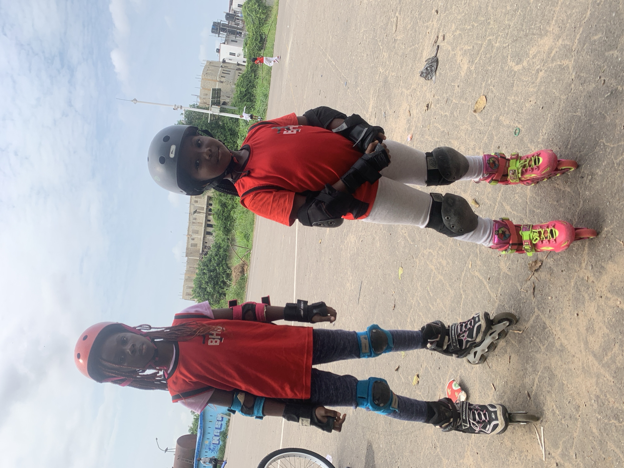 Two young girls are inline skating outdoors. The girl on the left is wearing a red vest over a pink t-shirt, blue and black elbow pads, black gloves, blue knee pads, grey leggings, and black and pink inline skates. She has dark braided hair under a red helmet. The girl on the right is wearing a red t-shirt, white leggings, black elbow pads, black gloves, grey knee pads, and pink and green inline skates. She has dark hair under a black helmet. They are on a paved surface, possibly a road, with some debris on the ground. In the background, there are buildings under construction, trees, and a cloudy sky. The scene appears to be in Alagbede, Nigeria, given the context.