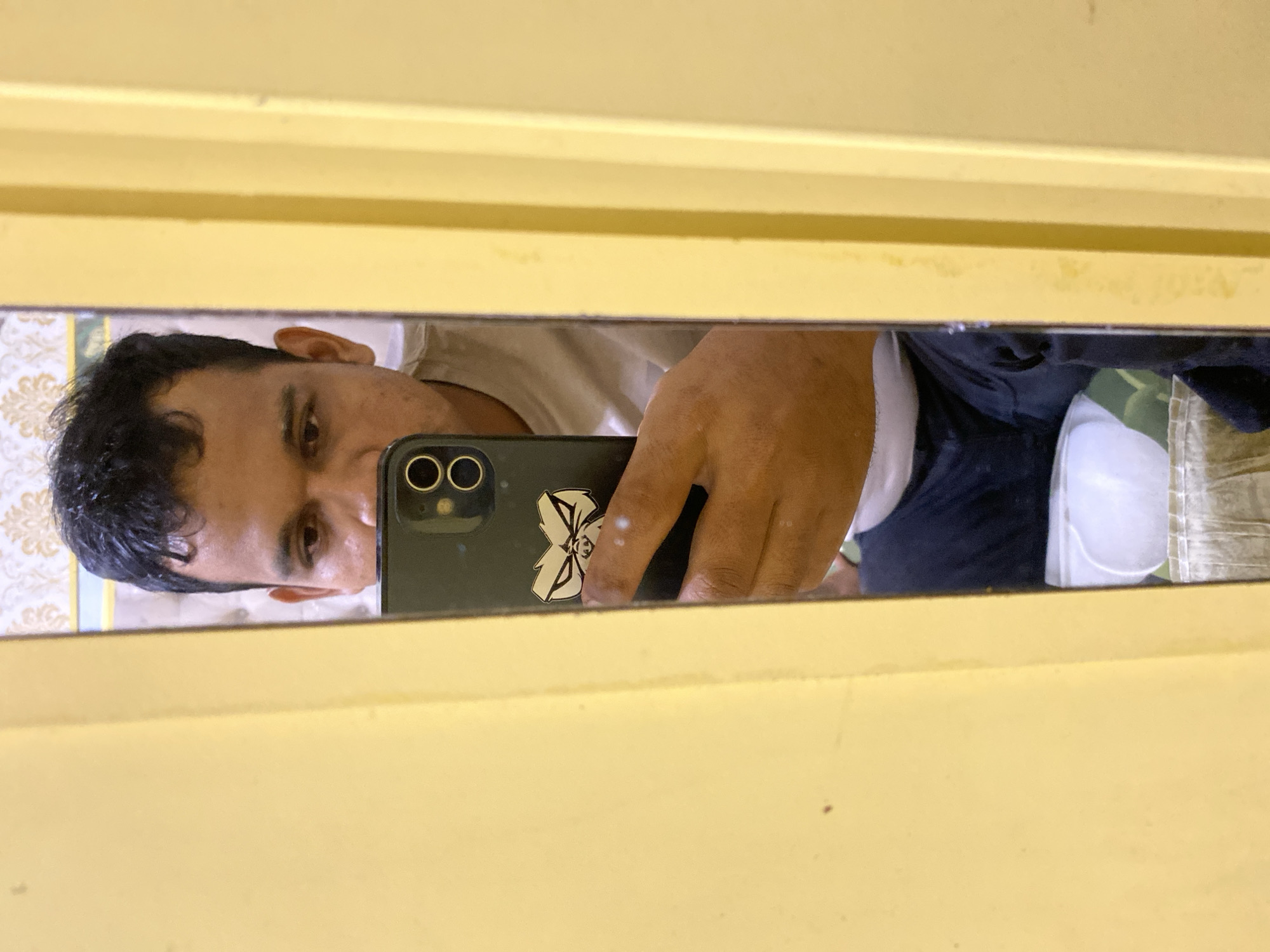 A medium shot captures a man taking a selfie in a full-length mirror. He is positioned to the left of the frame, his face and torso reflected. The mirror is framed by yellow door jambs. He is looking at his phone, which is held in his right hand, its screen facing him. The phone has a sticker of a cartoon character with large eyes and a stern expression on its back. The man is wearing a light-colored t-shirt and dark pants. Behind him, in the mirror's reflection, a white object, possibly a folded shirt or towel, is visible on a surface. The background of the room is out of focus, with a patterned wallpaper visible in the reflection. The lighting is dim, suggesting an indoor setting.