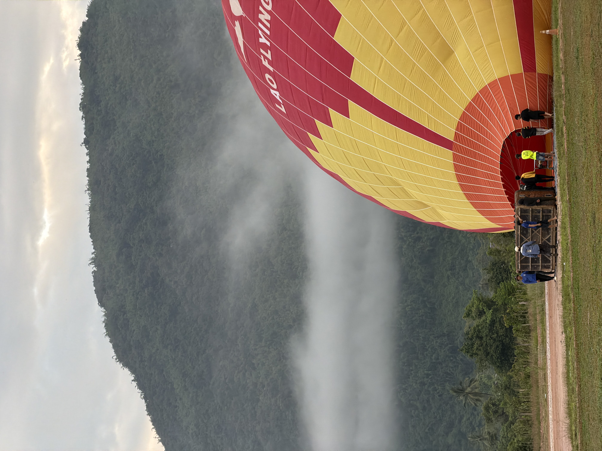 An aerial view of a landscape with a hot air balloon being prepared for flight. The balloon is a vibrant yellow and red, with the text "LAO FLYING" visible on its surface. Several people are gathered around the balloon, appearing to be part of the crew preparing for launch. The scene is set against a backdrop of lush green mountains, partially obscured by a layer of mist. The ground is covered in grass and dirt, with a dirt road leading to the balloon. In the distance, trees and a palm tree are visible. The sky is overcast with clouds, allowing some light to filter through. The location is the city of Vang Vieng, Laos, known for its stunning natural beauty and hot air balloon rides.