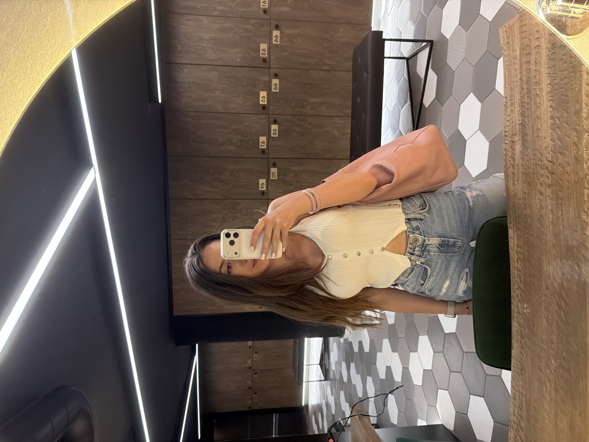 A woman stands in a locker room, taking a mirror selfie. She is wearing a white ribbed tank top and distressed denim shorts. She holds a pink handbag in her left arm and her phone in her right hand, which is capturing the reflection. The locker room has hexagonal tile flooring in gray and white, dark wood lockers, a black bench, and modern overhead lighting. The scene appears to be in Sofia, Bulgaria, suggesting a gym or fitness facility.
