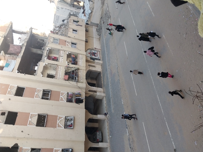The image shows a damaged multi-story building with significant structural damage. People are walking along a road next to the building. The scene suggests a conflict zone or aftermath of a disaster. The building has missing walls, exposed interiors, and visible debris. The people on the road appear to be civilians walking away from the damaged building. The overall atmosphere of the image is somber and suggests loss or destruction.