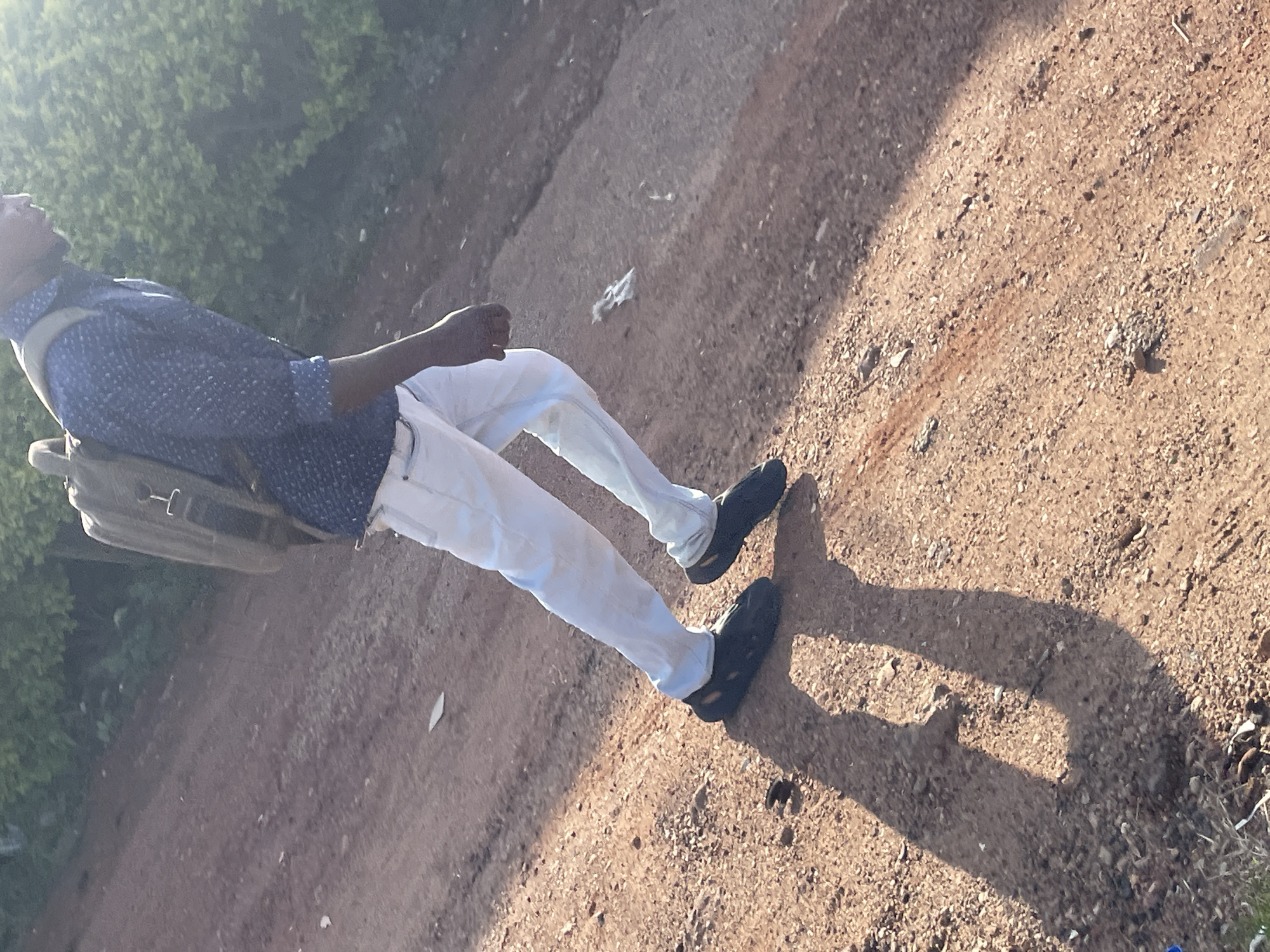 In Barakin Lerawi, Nigeria, a person walks uphill on a dusty, reddish-brown dirt path. The individual wears a blue, patterned button-down shirt, light-colored pants, and dark clogs. A backpack is slung over their shoulders. The ground is uneven and covered with small pebbles and dirt. Lush green foliage is visible in the background, with sunlight casting a strong shadow of the person on the path.