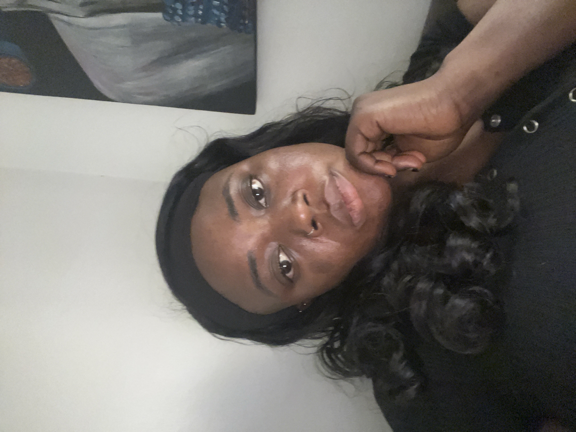 A medium shot photograph captures a Black woman posing against a white wall. The photograph has a slight tilt to the right. The woman is resting her chin in her hand, gazing directly into the camera with a serious expression. She has a black headband, and dark curly hair cascading around her face and shoulder. She is wearing a black shirt with metal ringlet details. A painting is seen in the top left corner of the image. The location of this image is unknown, and there is no information given to indicate that the image was taken in Abuja, Nigeria.