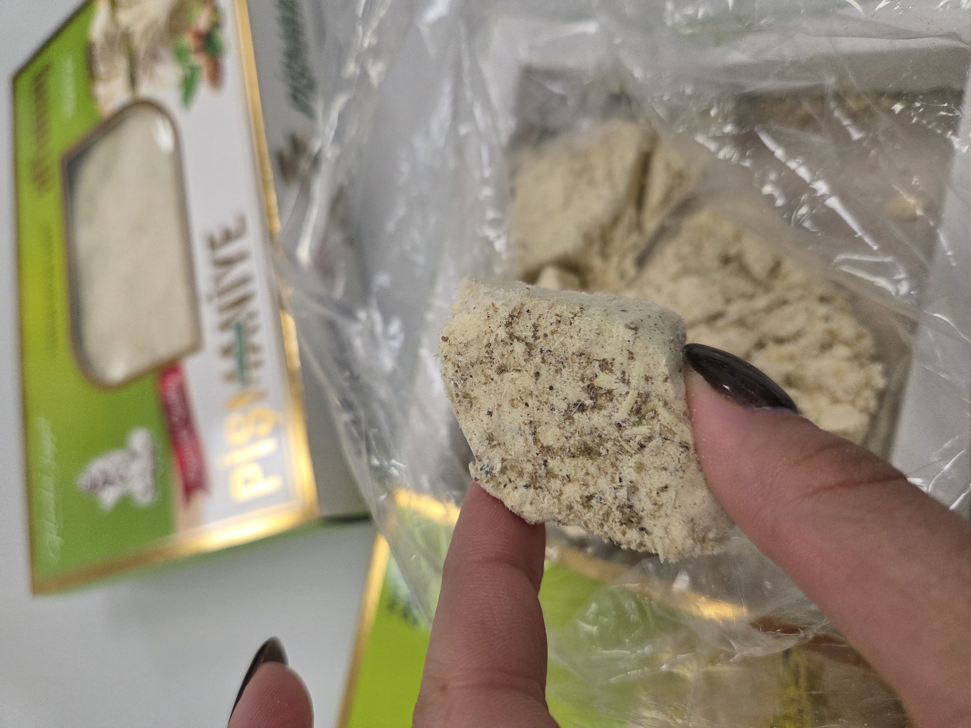 A close-up image shows a piece of halva held between a thumb and index finger. The halva is a light tan color with darker specks. A clear plastic bag partially contains more halva in the background. To the left, a green and gold box labeled "PİŞMANİYE" is visible.