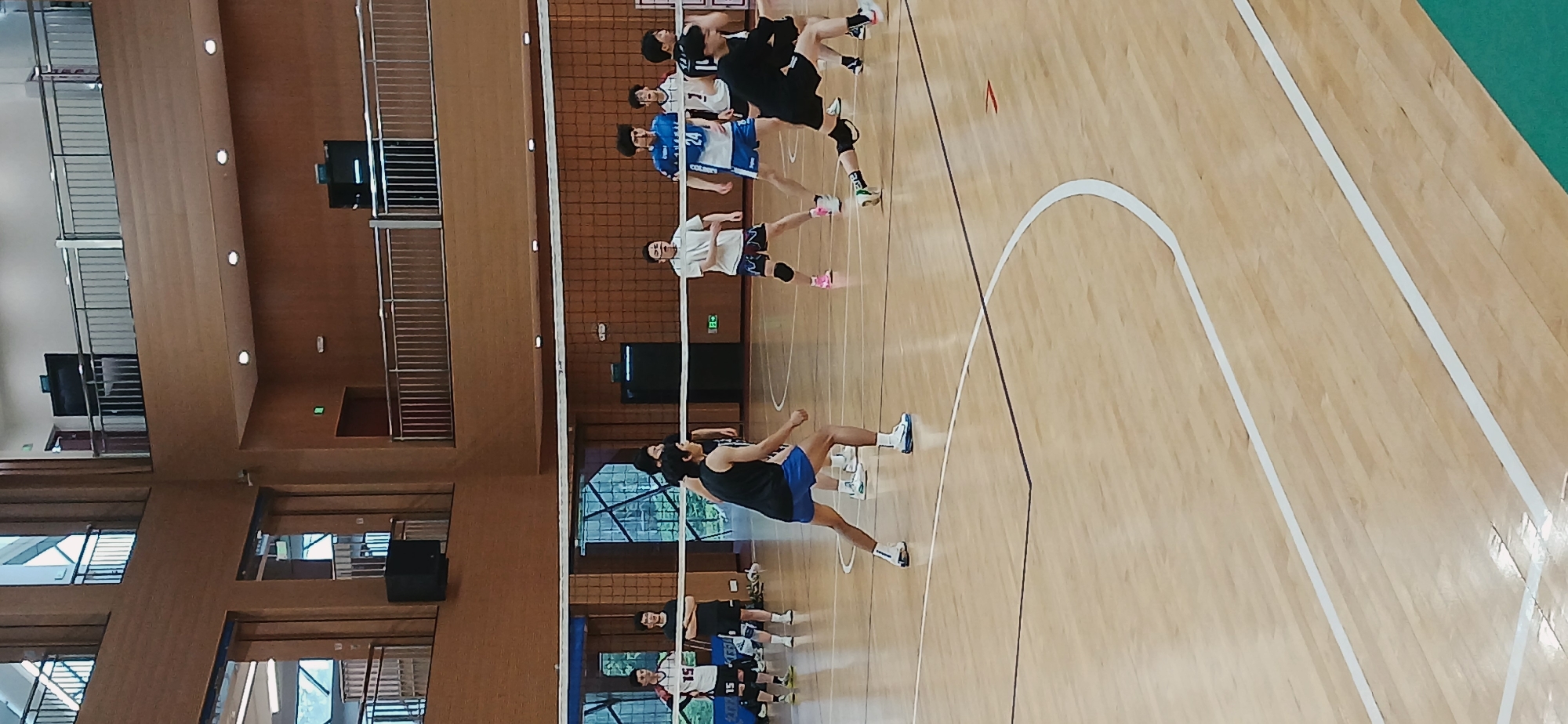 The image shows a volleyball game being played indoors on a wooden court. Several players are visible on both sides of the net, in volleyball attire. The environment appears to be a gymnasium or indoor sports facility. There is a railing system visible on the walls to the left of the volleyball net and a basketball hoop on the right corner of the image.
Given the context provided (Chongqing High-tech Zone, China), the location is a sports facility within the High-tech Zone.