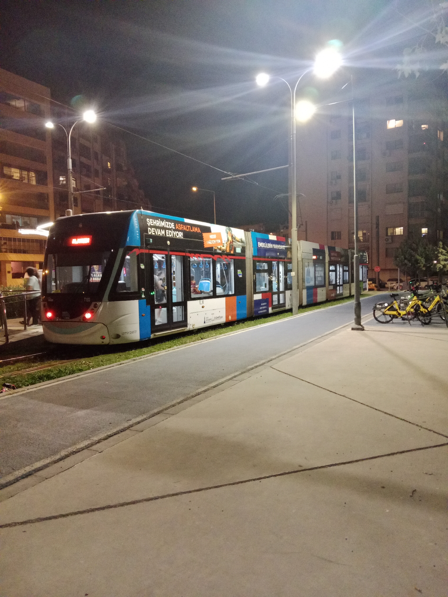 In the city of Karşıyaka, Turkey, at night, a modern tram with a vibrant advertisement wraps its side, is stopped at a station. The tram's exterior displays text in Turkish, including "ŞEHRİMİZDE ASFALTAMA DEVAM EDİYOR" and "EMEKİERİN YANINDAYIZ." The station platform is paved with concrete and bordered by a grassy strip. A person stands on the platform, partially obscured. In the background, multistory apartment buildings are visible, their windows illuminated. Streetlights cast a bright glow over the scene. To the right of the tram, two yellow rental bicycles are parked on the sidewalk. The overall impression is one of urban transit infrastructure at night, with commercial messaging integrated into the public space.