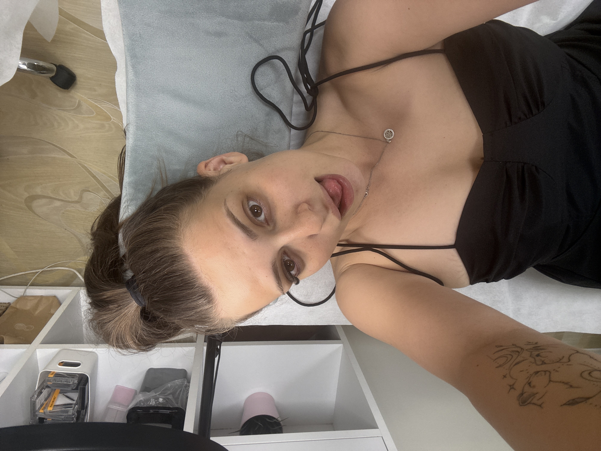 A young woman with her hair in a high ponytail and wearing a black spaghetti strap dress is lying down, taking a selfie. She is sticking her tongue out and looking at the camera. She is wearing a silver necklace with a circular pendant. There is a tattoo of animals and celestial elements on her left arm. In the background, there are shelves filled with various items, including what appears to be beauty products and a hairdryer. The setting looks like a beauty salon or a personal grooming area. The floor has a wood-like pattern. This scene is not specific to the city of Plovdiv, Bulgaria, as there are no discernible landmarks or indicators of that location.
