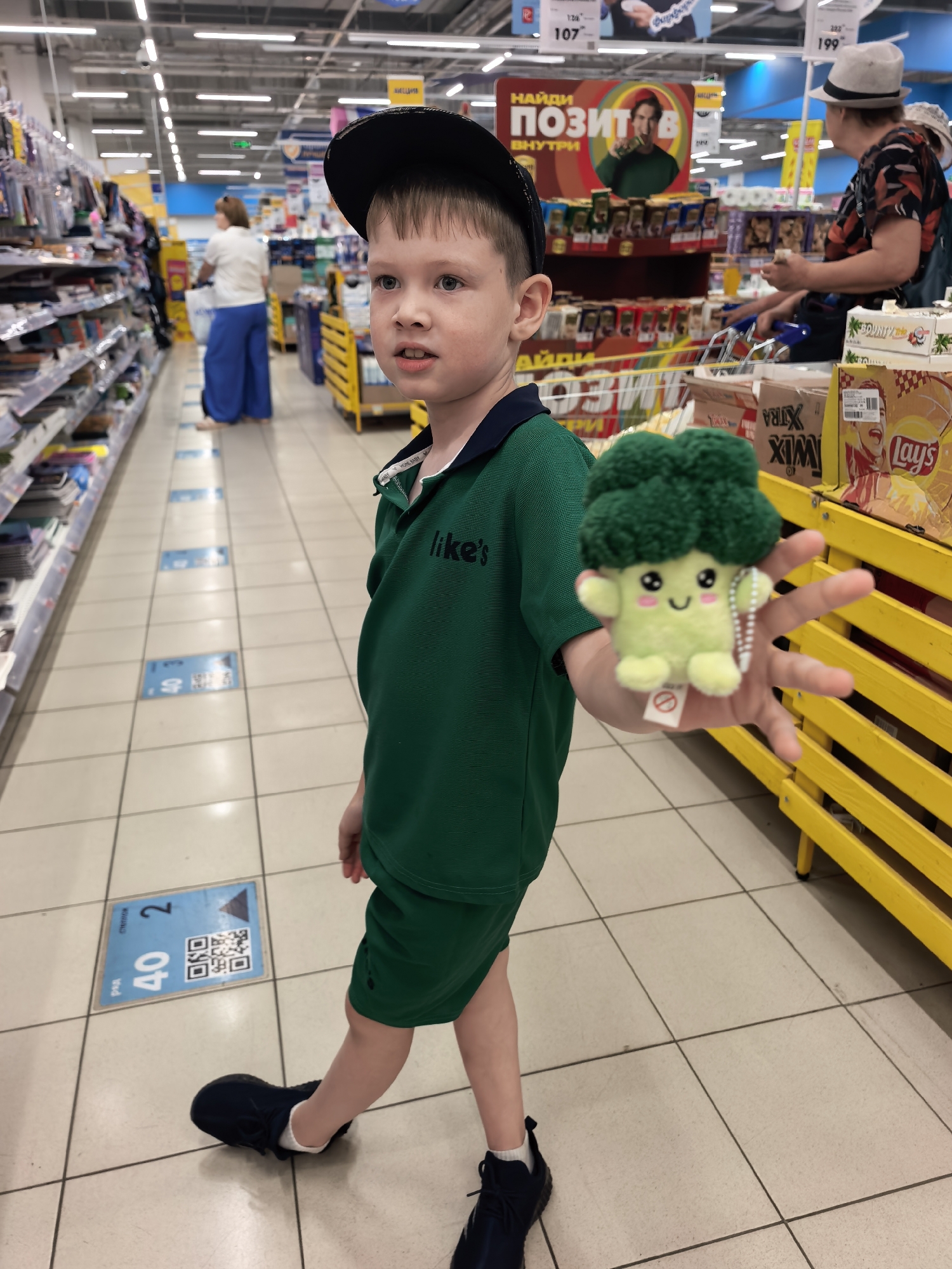 A young boy wearing a black baseball cap and a green polo shirt and shorts stands in a grocery store aisle, holding a plush toy broccoli. He is looking towards the left of the frame. The aisle is well-lit with fluorescent lights, and shelves stocked with various products are visible on both sides. In the background, other shoppers can be seen browsing. The floor is tiled, and there are blue markers on the floor indicating rows and numbers. The overall setting appears to be a typical supermarket in the city of Sterlitamak, Russia.