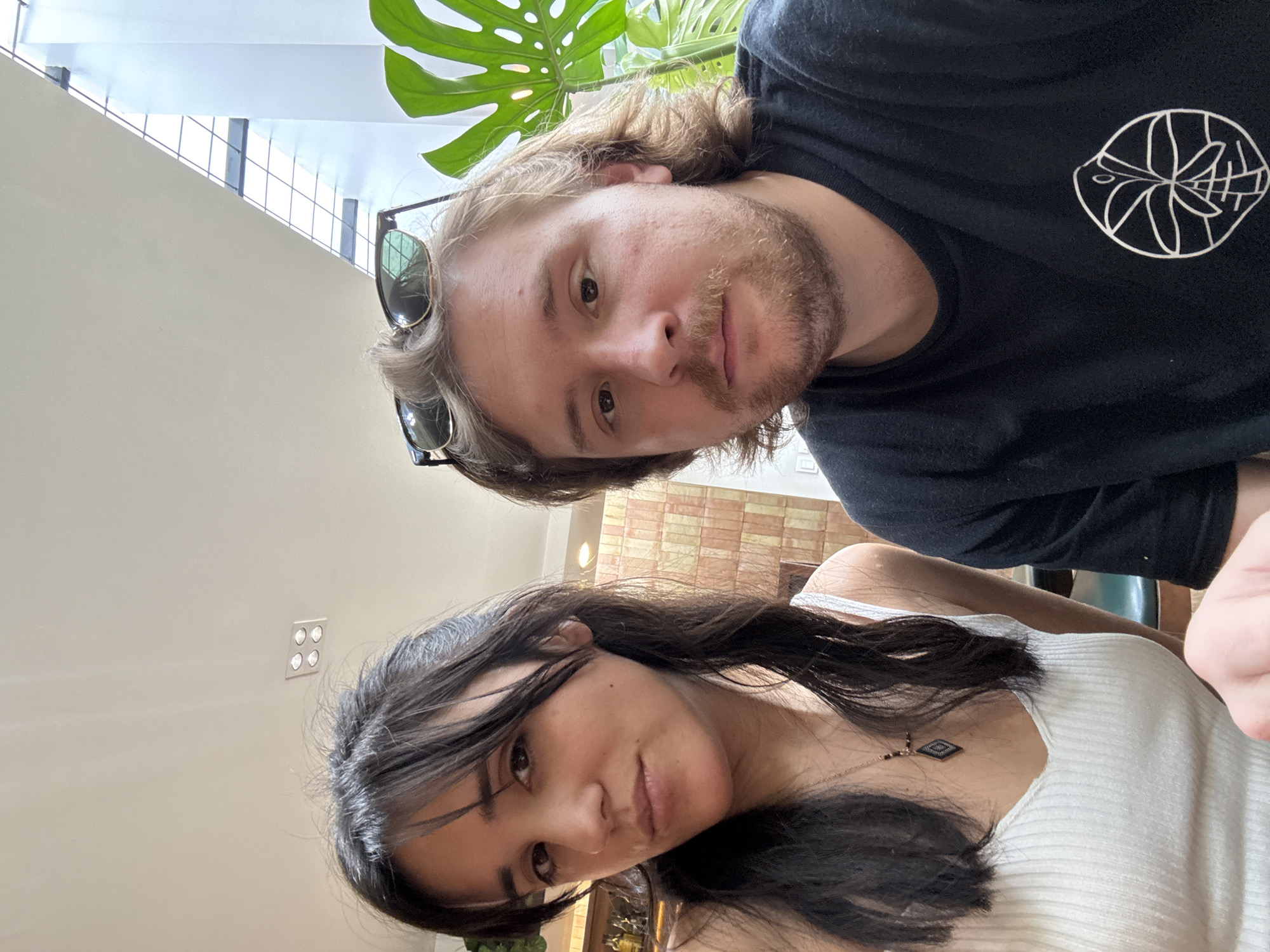 Two people, a man and a woman, are taking a selfie indoors. The man is wearing a black t-shirt with a white circular design on the chest and has sunglasses on his head. He has light brown hair and a beard. The woman is wearing a white tank top and a necklace with a diamond-shaped pendant. She has dark hair with bangs. A large Monstera plant is visible in the background, along with a staircase and a brick wall. Based on the interior, it could be a cafe or restaurant setting in Cuautitlán Izcalli, Mexico.