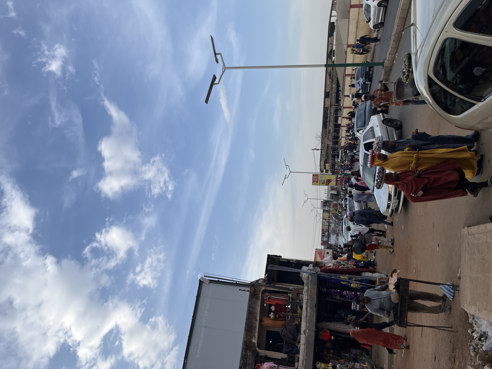 This image depicts a bustling street scene in Jos, Nigeria, under a bright blue sky dotted with scattered white clouds. The perspective captures a busy commercial or market area, teeming with people and activity.
On the left side of the frame, several buildings house open-front shops or market stalls, displaying various goods, including clothing. A large white signboard on one building shows inverted text, partially decipherable as "AVIA TUMEB AS ROV DEVOO," likely an advertisement or shop name. In front of these establishments, people are visible, including a man operating a charcoal grill or cooking stand, and another individual in red walking by.
The central and right portions of the image showcase the street and sidewalk, crowded with pedestrians and vehicles. Several white cars are parked along the road, suggesting either heavy traffic or a designated market parking area. Numerous people are seen walking, standing, and interacting. Notable are vendors, one pushing a cart loaded with large fruits (possibly watermelons), and others carrying trays of eggs balanced on their heads.
Tall, modern streetlights, equipped with double solar panels, punctuate the clear sky. The bright daylight and lack of harsh shadows indicate it is likely midday or early afternoon. The scene vibrantly portrays the daily commerce and movement characteristic of an urban market in the region.