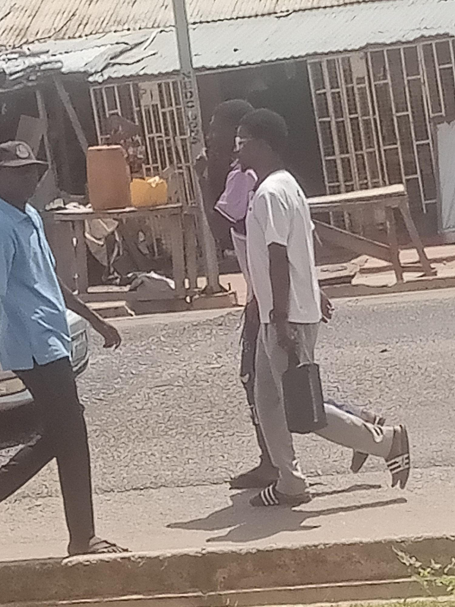 In Jalingo, Nigeria, three individuals walk along a paved area adjacent to a street. The person on the far left, wearing a light blue collared shirt and dark pants, walks away from the camera. In the center, a person wearing a purple shirt and dark pants walks beside another individual. This second person, on the right, is wearing a white t-shirt and grey sweatpants, carrying a dark rectangular bag. Behind the individuals, a building with a corrugated metal roof and barred windows is visible, along with wooden tables and barrels. The ground is covered in loose gravel and debris.