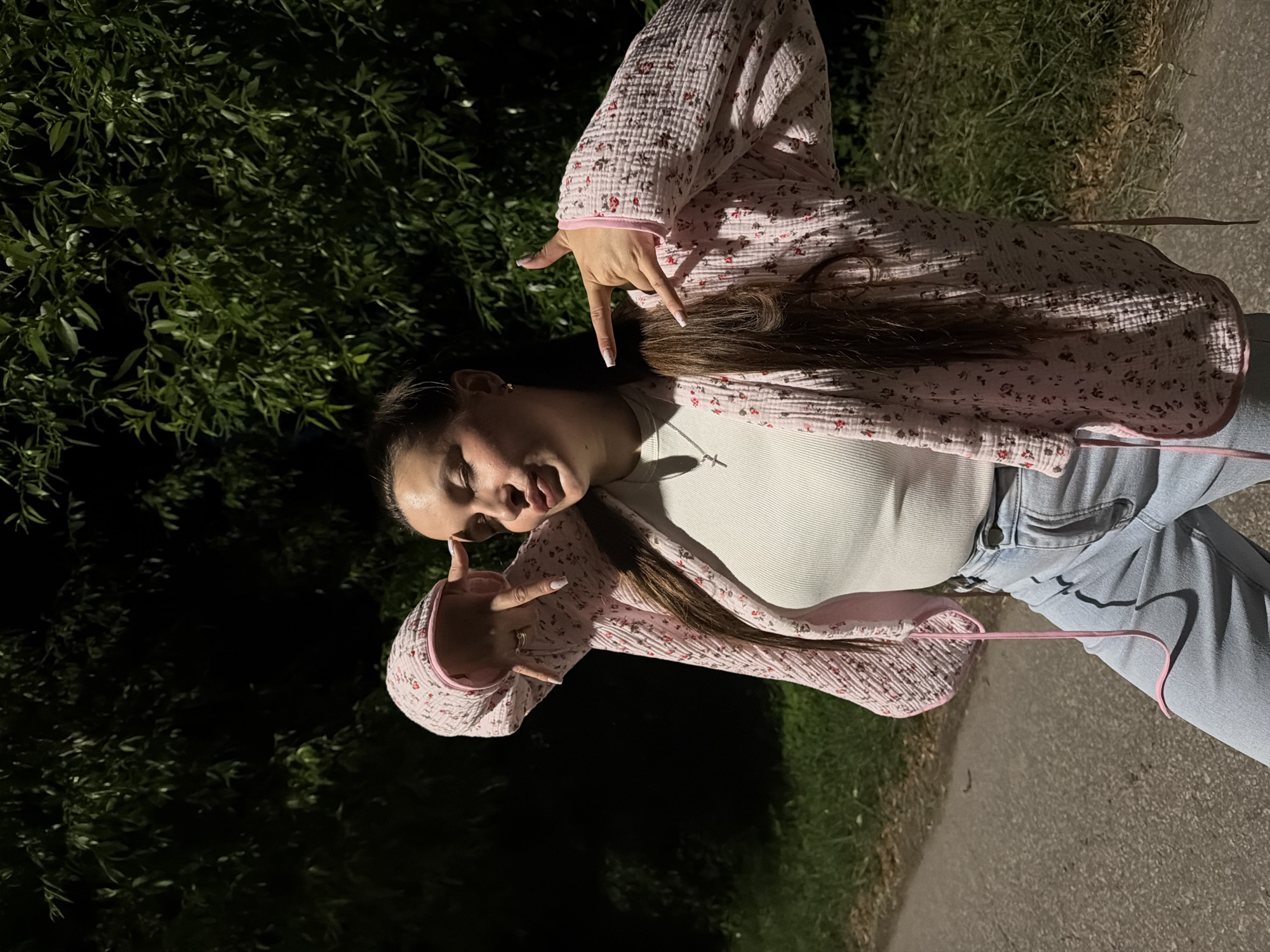 A young woman with long brown hair is posing for a photo outdoors at night. She is wearing a white ribbed tank top, light blue jeans, and a pink floral patterned open-front cardigan. She has her eyes closed and her tongue sticking out slightly, while holding up her hands in a gesture that could be interpreted as horns or a peace sign. The background consists of dark foliage and a paved surface, likely a road or path. The scene is illuminated by artificial light, casting shadows. The location is specified as the city of Krasnyy Mayak, Russia.