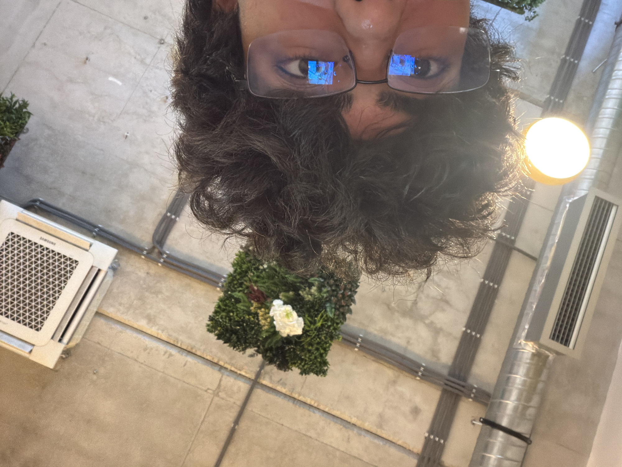 An indoor shot, taken from above and facing directly down, shows a person wearing glasses with a light-reflective indoor scene within the lenses, with dark curly hair, and an upside-down view of the ceiling. The ceiling shows a square green floral decoration with a white flower, a Samsung-branded ceiling air-conditioning unit, exposed pipes, metal ductwork, a recessed rectangular air vent, and a round light fixture. The photo seems to be taken indoors, but there are no clear landmarks that would associate it with a location in Podgorica, Montenegro.