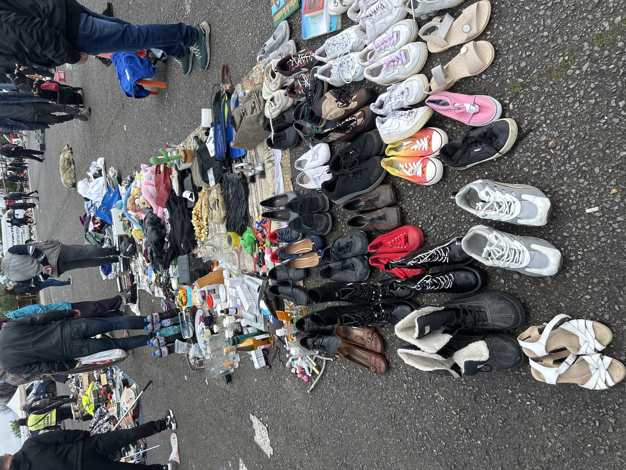 The image shows a flea market in Sofia, Bulgaria. Items for sale are laid out on the pavement, including a wide assortment of shoes (sneakers, sandals, boots), books, toys, glassware, and other miscellaneous household items. Several people are standing around, presumably browsing or interacting with the vendors. The ground is asphalt.