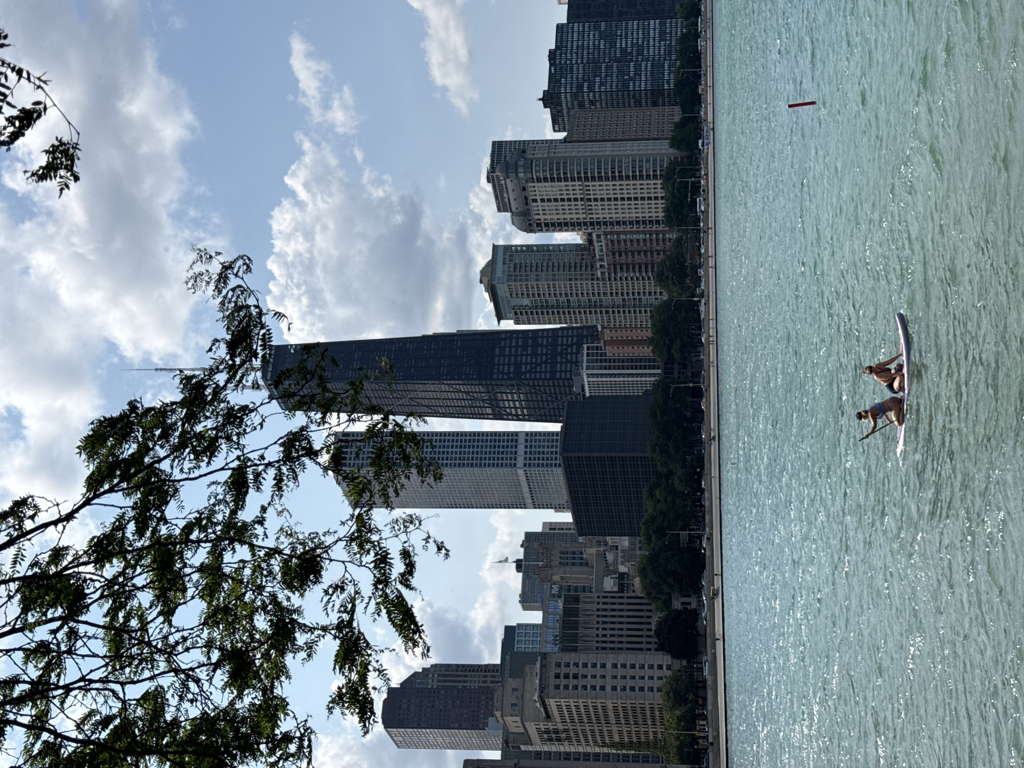Two individuals are paddleboarding on a body of water with a city skyline in the background. The paddleboarders are wearing swimwear. One person is seated on the board, holding a paddle. The other person is also seated on the board. The water is blue-green and appears to be choppy, with sunlight reflecting off the surface. The city skyline features numerous tall buildings, including a very tall, dark skyscraper. Trees line the waterfront between the buildings and the water. The sky is blue with scattered white clouds. A red marker pole is visible in the water to the right of the paddleboarders. The scene is located on the Lake Michigan waterfront in Chicago, Illinois.