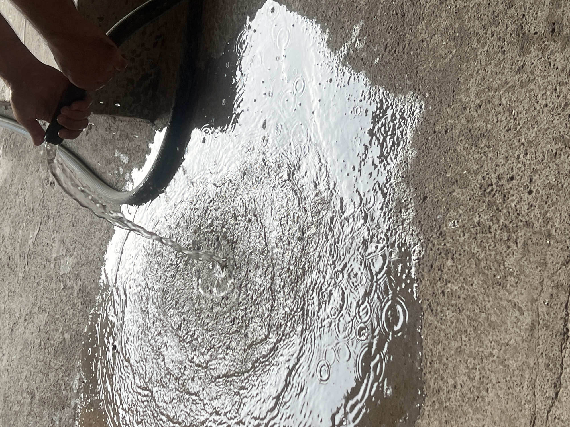 A close-up, overhead shot shows a stream of water being sprayed from a black hose nozzle held by a person's hands. The water lands on a textured concrete surface, creating a large puddle with ripples and circular patterns. The water is clear and reflects light. The concrete appears to be outdoors, possibly a driveway or patio. The focus is on the water and the hose, with the hands and the concrete serving as background elements.