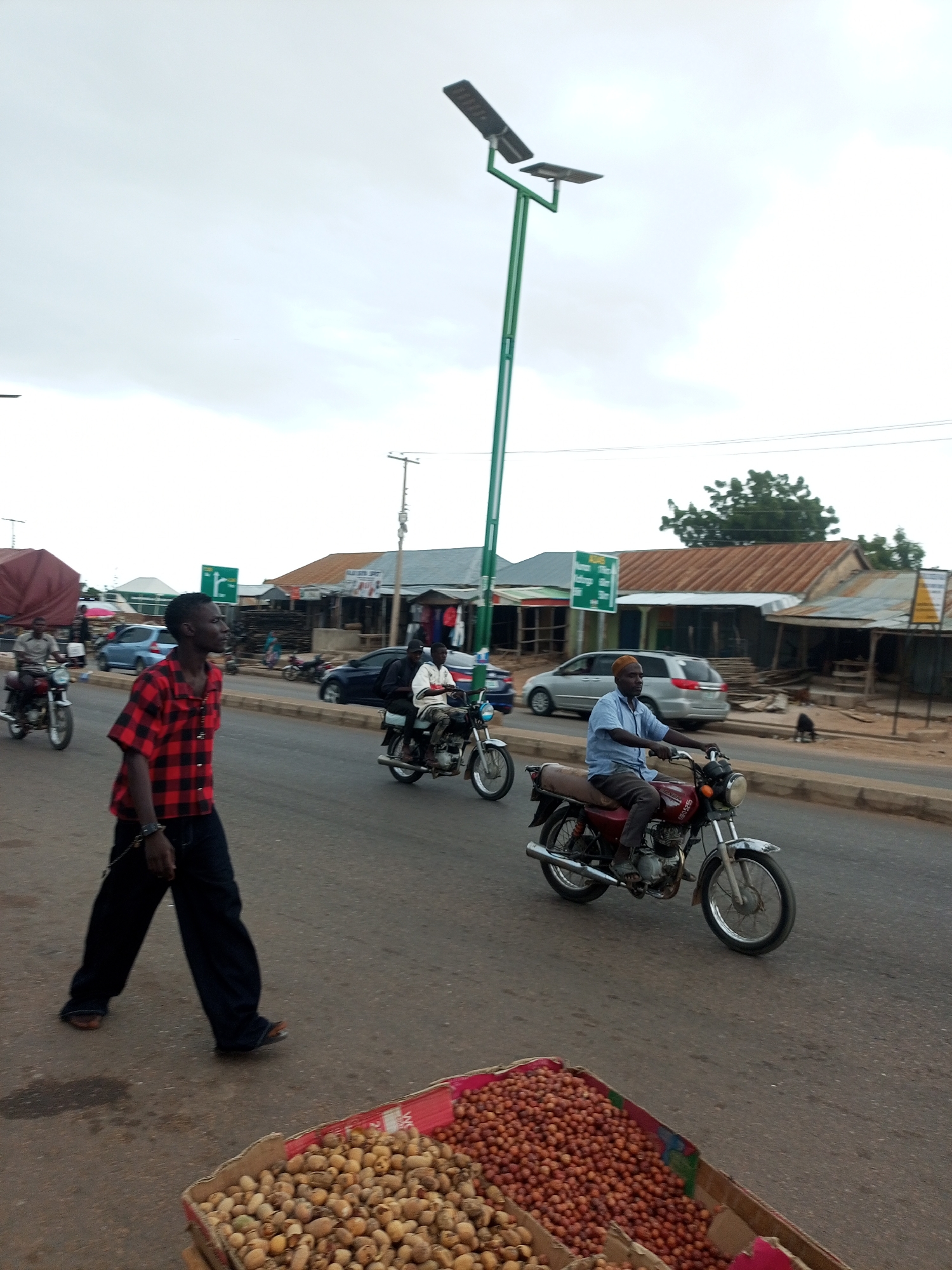 In Tempure, Nigeria, a street scene unfolds under an overcast sky. A tall, green street lamp with two solar panels stands prominently on the right side of the road. The street is paved, with a lighter colored curb on the right. In the foreground, to the left, a young man walks away from the camera, his body angled slightly to the right. He is wearing a red and black checkered shirt and dark, loose-fitting pants. His right hand is extended, and a black chain is visible connecting his wrist to something out of frame.
Further down the street, several motorcycles are present. One is traveling towards the camera on the left, ridden by a person in a light-colored shirt. Another motorcycle is moving away from the camera in the middle lane, with two individuals on it, one in a white shirt and the other in a darker top. To the right, a man wearing a light blue shirt and dark pants, with a light-colored cap, rides a red motorcycle. He is looking forward.
The background features buildings with corrugated metal roofs, suggesting a market or commercial area. Some of these buildings appear to be shops, with goods displayed outside. A silver minivan is parked or moving in the background. A few trees are visible in the distance. In the bottom right corner of the image, a cardboard box filled with small, light-brown nuts is visible on the ground. The overall atmosphere is that of a busy, everyday street in a Nigerian city.