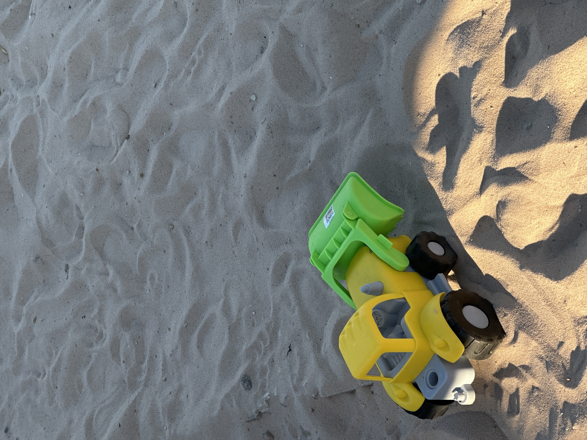 The image shows a yellow and green plastic toy truck on a sandy surface, possibly a beach. The truck is positioned on the right side of the frame, with its green bucket raised. The sand appears textured and rippled, with variations in color due to light and shadow. In the upper right, there are shadows cast onto the sand, suggesting the presence of trees or other objects blocking the sunlight. The location is not identified in the image.