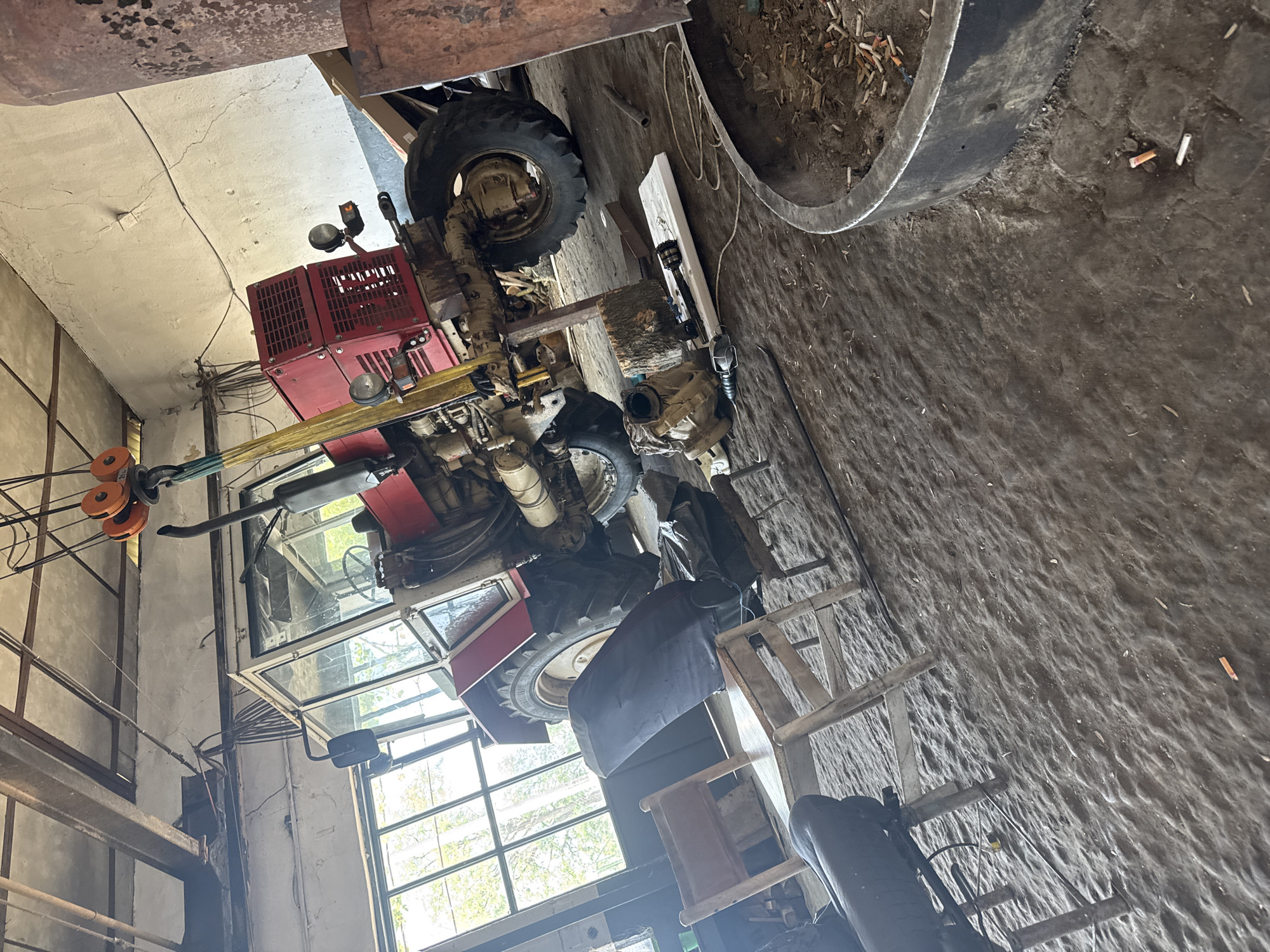 The image captures an indoor scene, likely a workshop or barn in Sarnevo, Bulgaria, featuring a red tractor positioned on its side. The tractor has large tires, a red engine block with grated covers, headlights, and a cabin with glass windows, suggesting it is either undergoing significant repair or is stored in this unusual orientation.
The surrounding environment is functional and somewhat cluttered. The floor is an uneven, textured surface of dirt or rough concrete, with various items scattered across it. These include wooden planks, a log, and a tan-colored mechanical component that appears to be a gearbox or differential. A prominent dark, circular metal container, possibly a trough or large basin, is filled with dirt, wood shavings, and numerous cigarette butts. Several cigarette butts are also visible directly on the floor.
Natural light streams into the space through large windows in the background, indicating it is daytime. Overhead, a hoist or pulley system with orange wheels, along with various cables and wires, suggests the capacity for lifting heavy objects. The walls are plain, possibly concrete, and show some cracks and utility lines. A wooden ladder-like structure and a black tarp or cover draped over an object are also present.
No people are visible in the image. The scene conveys an atmosphere of ongoing work or preparation for maintenance on the tractor within an industrious, rural setting. No legible text is visible in the media file.