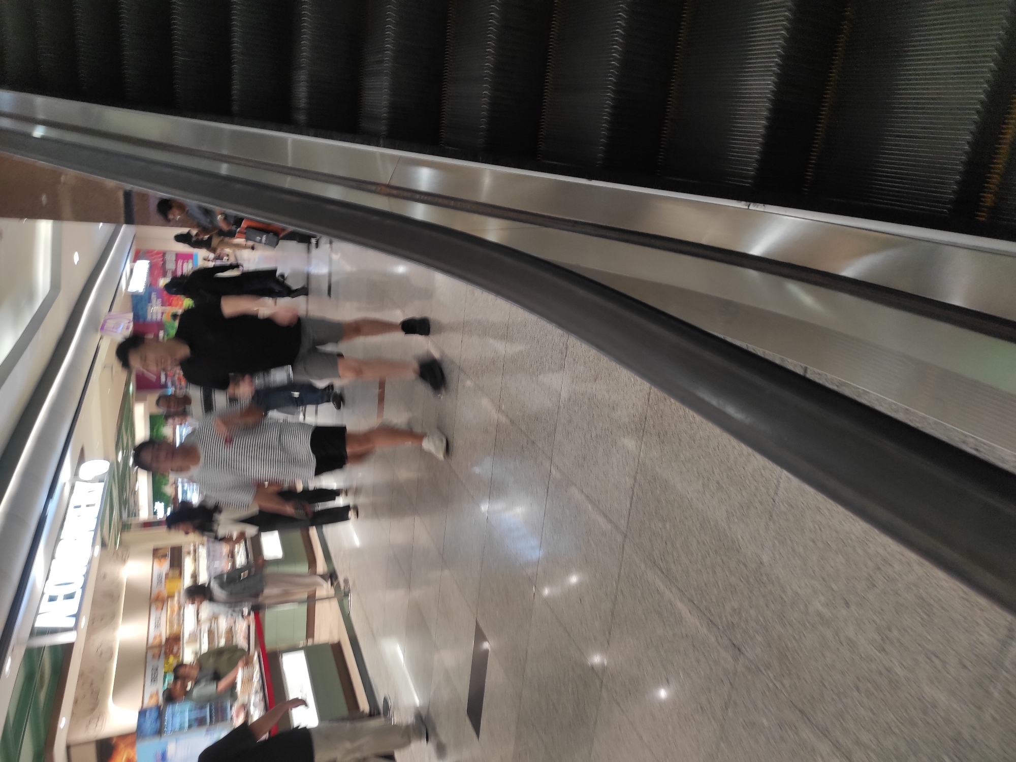 The image shows an indoor escalator in a commercial setting, likely a shopping mall. The escalator is on the right side of the frame and is dark gray with silver accents. The steps of the escalator are clearly visible. In the background, there are people walking and shopping. One prominent sign reads "INFOBANKERY," suggesting a bakery or food establishment. The floor is tiled and appears to be made of granite or a similar material. The lighting is bright and overhead, typical of a commercial space. The overall scene depicts a busy commercial area with shoppers utilizing the escalator. The image was taken at Tunjungan Plaza 1, Surabaya, Indonesia.