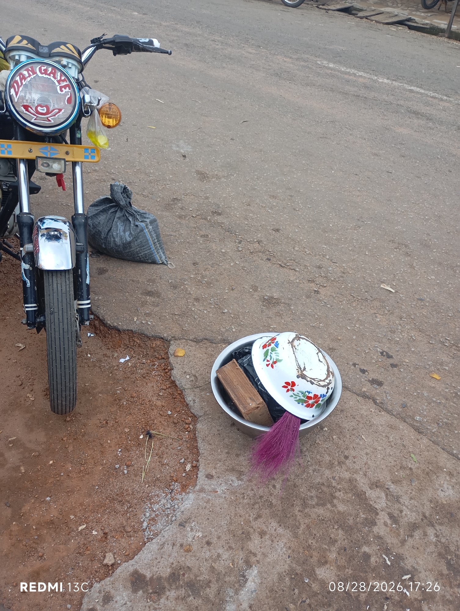 This is a photograph of a motorcycle parked on the side of a road in Shendam, Nigeria. The motorcycle is black and yellow with chrome accents. The front wheel is visible, and the headlight has a decal that reads "DAN CAYE". A black bag is placed on the ground behind the motorcycle. To the right of the motorcycle, a metal bowl contains a wooden object, a black bag, and a purple brush. The ground is a mix of dirt and asphalt. The date stamp indicates the photo was taken on August 28, 2026, at 17:26.