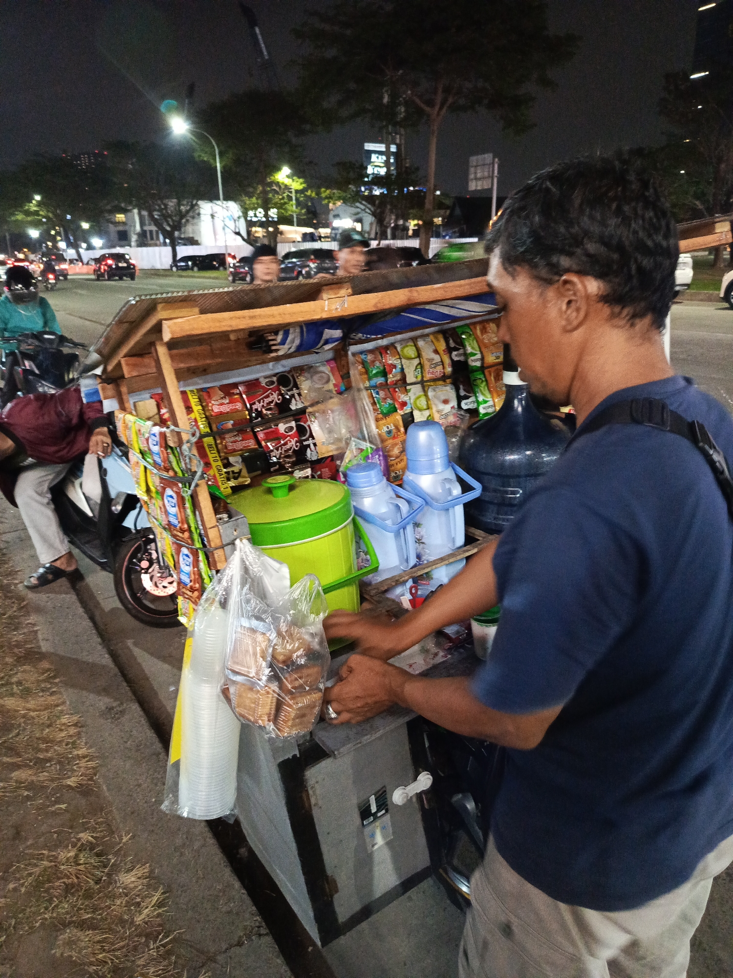 The image depicts a street scene at night in Tangerang, Indonesia, likely in a commercial or residential area given the presence of streetlights and buildings in the background. In the foreground, a man in a dark blue t-shirt and light-colored shorts is standing behind a mobile food cart. The cart is wooden and appears to be mounted on a motorcycle, with a canopy of wood and fabric. It is laden with various items, including bags of snacks, bottled drinks, and two large water jugs, one dark blue and the other a light blue dispenser. A green cooler is also visible. In front of the cart, a stack of clear plastic cups and a bag of biscuits are arranged. To the left of the cart, a woman wearing a turquoise jacket and dark pants is seated on a motorcycle, with her hands resting on the cart. The street is paved, and dry grass is visible along the roadside. In the background, cars are driving by, and streetlights illuminate the scene, casting long shadows. Buildings, some appearing to be multi-story, and trees are also part of the environment. The overall atmosphere suggests a typical night scene of street vending in an urban Indonesian setting.