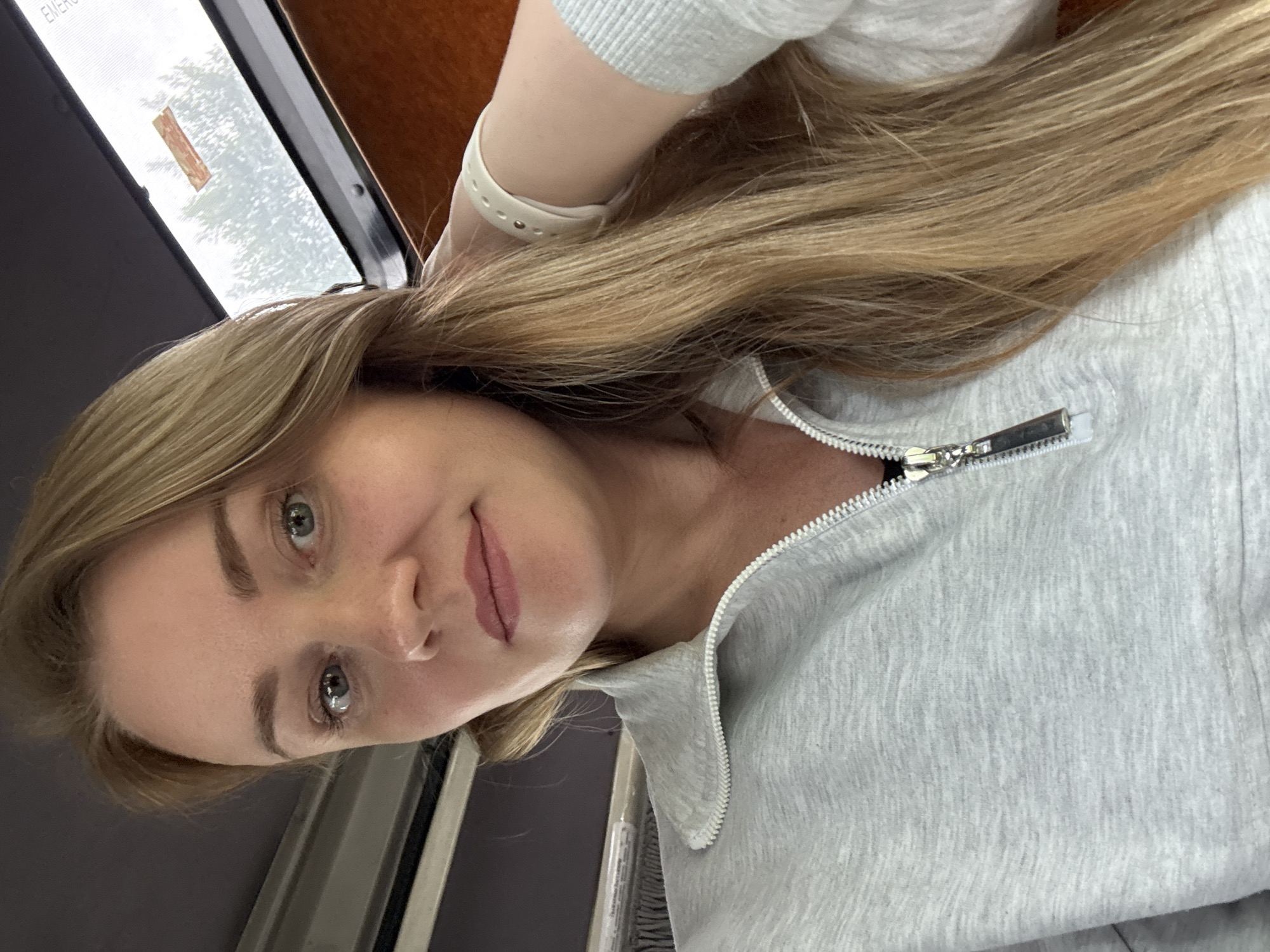 A female subject, appearing to be an adult, is captured in a close-up portrait, looking directly at the camera. She has long, straight blonde or light brown hair that falls over her shoulders. Her eyes are light-colored, and she displays a neutral facial expression with slightly parted lips. She is wearing a light gray quarter-zip sweatshirt with a silver-toned zipper. A white band, consistent with a smartwatch or fitness tracker, is visible on her left wrist. The background on the left is a dark, possibly padded, interior surface, while the upper right features a window looking out onto blurred green foliage, indicative of trees or bushes, under what appears to be an overcast sky. A small orange rectangular label is affixed to the window pane, and the word "EMERGENCY" is partially visible in the upper right corner of the window frame. Below the window on the right is an orange-brown textured surface, suggestive of interior paneling, possibly within a vehicle or small room. The overall context indicates the scene is located in the city of Chaikovsky, Russia.