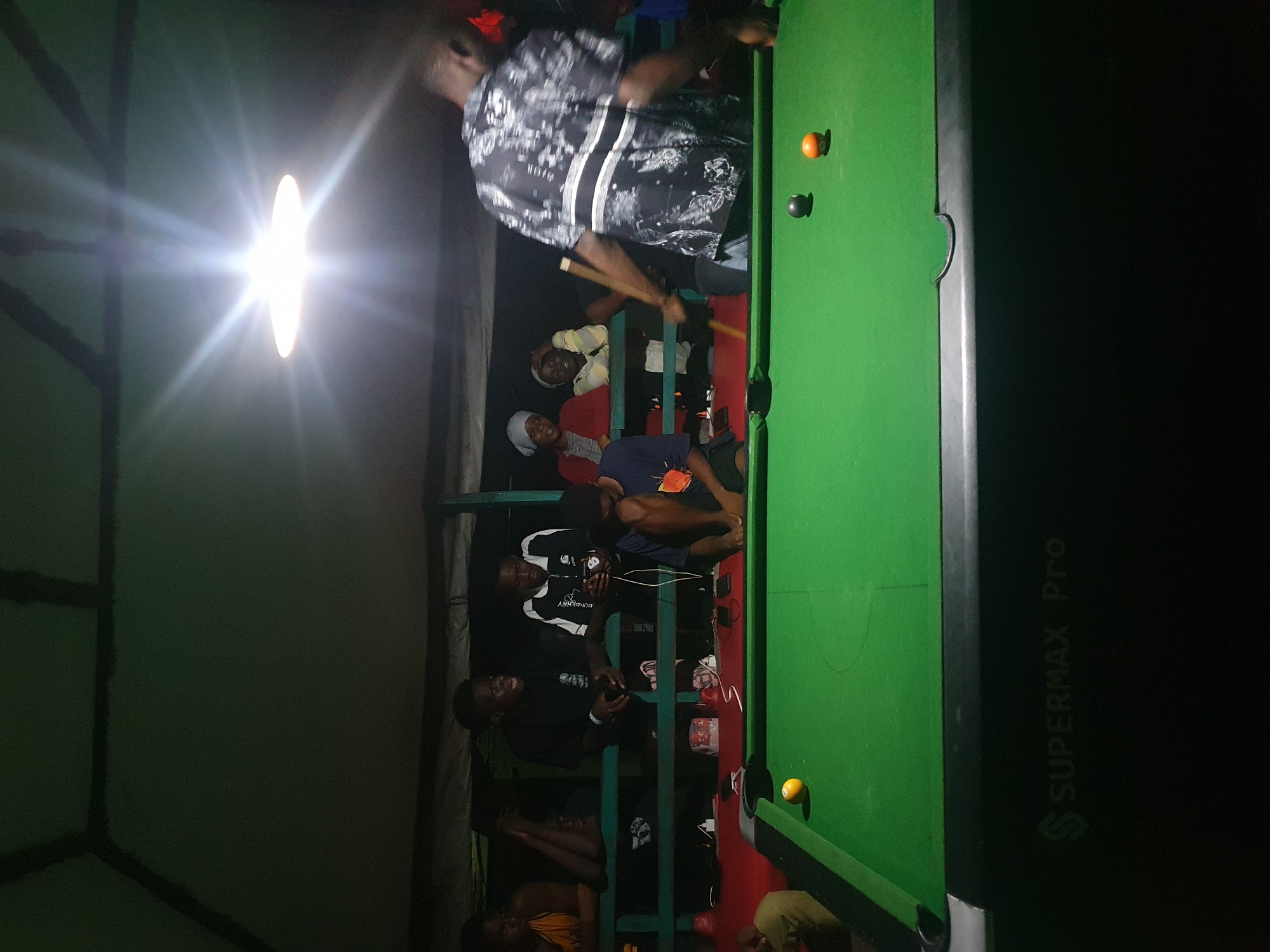 The image depicts an indoor pool hall in Lokoja, Nigeria, with a group of young men gathered around a green pool table. The lighting is dim, with a bright overhead light casting strong shadows. Several individuals are visible, some leaning on a railing, others standing or sitting near the table. One person is actively playing pool, holding a cue stick. Various pool balls are scattered on the table. The environment appears to be a casual gathering space. The pool table has "SUPERMAX Pro" branding on its side.