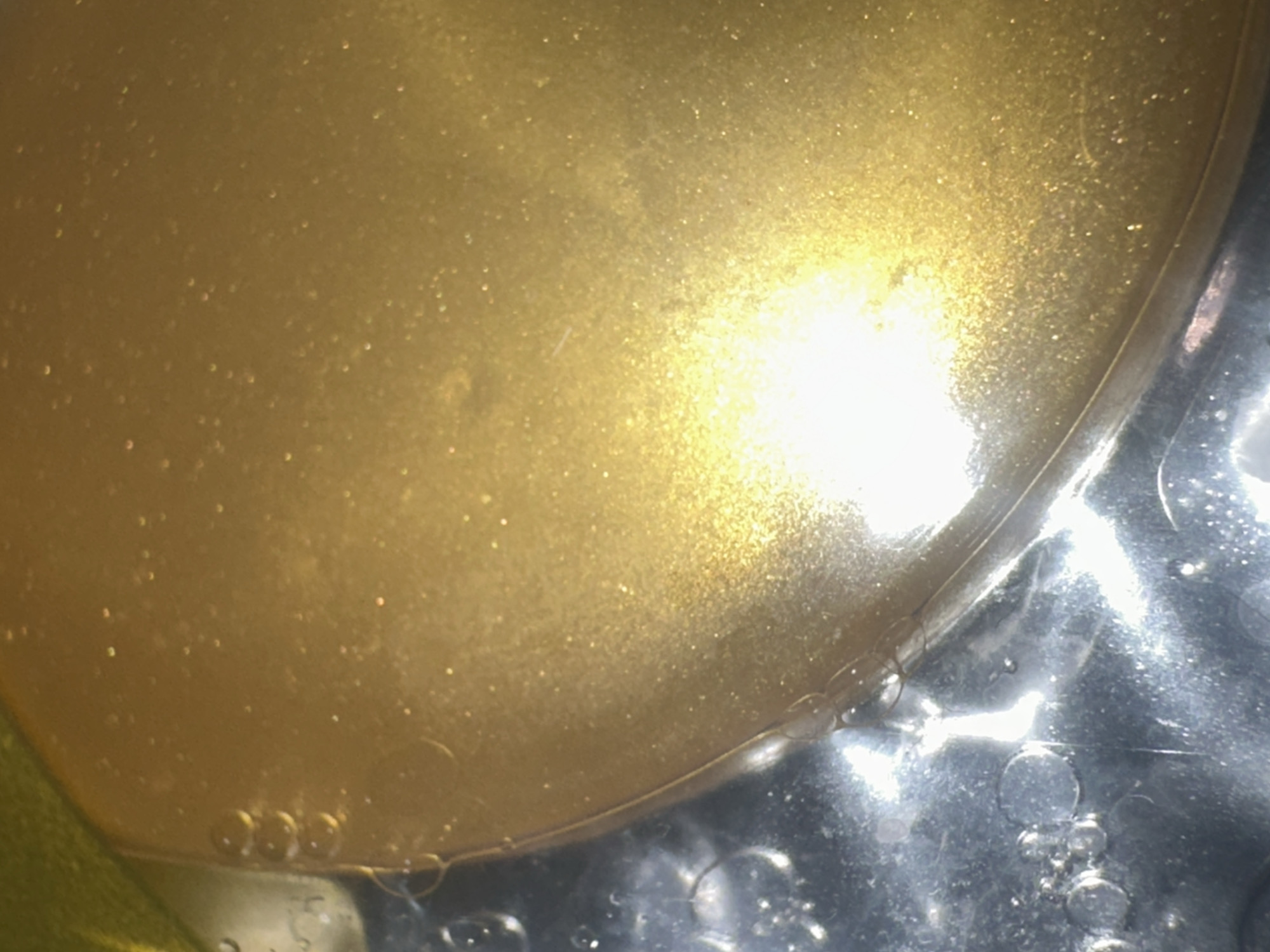 The image is a close-up, macro shot of a golden, viscous liquid, possibly oil or a cosmetic product, with fine shimmering particles suspended within it. The liquid has a smooth, glossy surface that reflects light intensely, creating a bright, hazy area in the center and lower right portion of the frame. The left side of the image shows a transparent container or surface with several small, spherical bubbles. A thin, curved line, which could be a reflection or an edge of the container, separates the golden liquid from the bubbly area. The background is out of focus and appears to be a dark surface.
There is no identifiable information or context within the image that would allow for its location in Letterkenny, Ireland, or any other specific city. The image focuses solely on the texture and reflective properties of the liquid and bubbles.