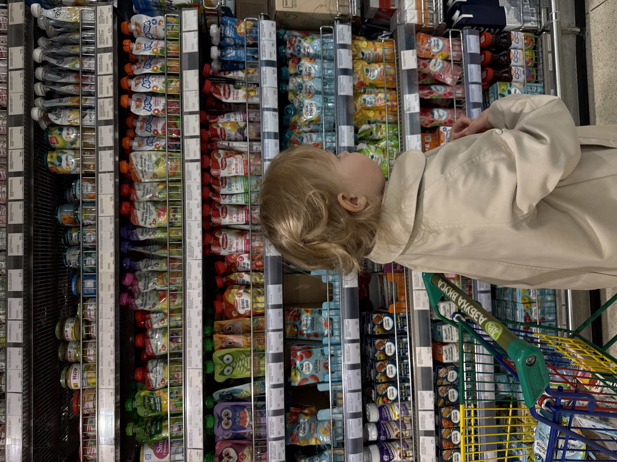 The image shows a young child in a supermarket aisle, viewed from a slightly elevated angle looking down. The child, with blonde hair, is wearing a light-colored jacket and is looking upwards at the products displayed on the shelves. A green and yellow shopping cart is visible to the right of the child, with the handle clearly showing "АЗБУКА ВКУСА" (Azbuka Vkusa), a Russian supermarket chain.
The shelves are fully stocked with a variety of baby food products, primarily in pouches and jars. Brands such as "Няня" (Nanny), "Semper", "HiPP", "ФрутоНяня" (FrutoNanny), and "Агуша" (Agusha) are visible. Product descriptions, mostly in Cyrillic, indicate items like "ЯБЛОКО, БАНАН" (Apple, Banana), "ПЕЧЕНЬЕ" (Cookies), "КУСОЧКИ" (Pieces), "КАША" (Porridge), and various fruit and vegetable purees. Prices, displayed in rubles (e.g., 249, 75.90, 169), are visible on small tags below each product. The overall setting is a brightly lit indoor commercial space.