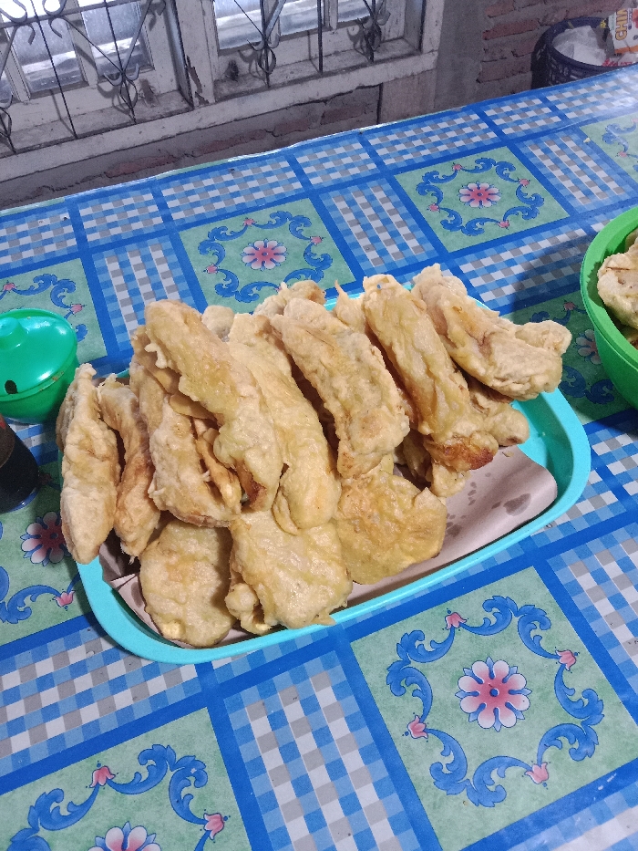 A pile of fried bananas sits on a light blue tray lined with paper. The bananas are golden brown and arranged densely. Adjacent to the tray, on the left, is a small green container. To the right of the tray, a green bowl partially filled with more fried food is visible. The setting includes a table covered with a blue and green patterned tablecloth. In the background, a window and a container add to the scene's context.