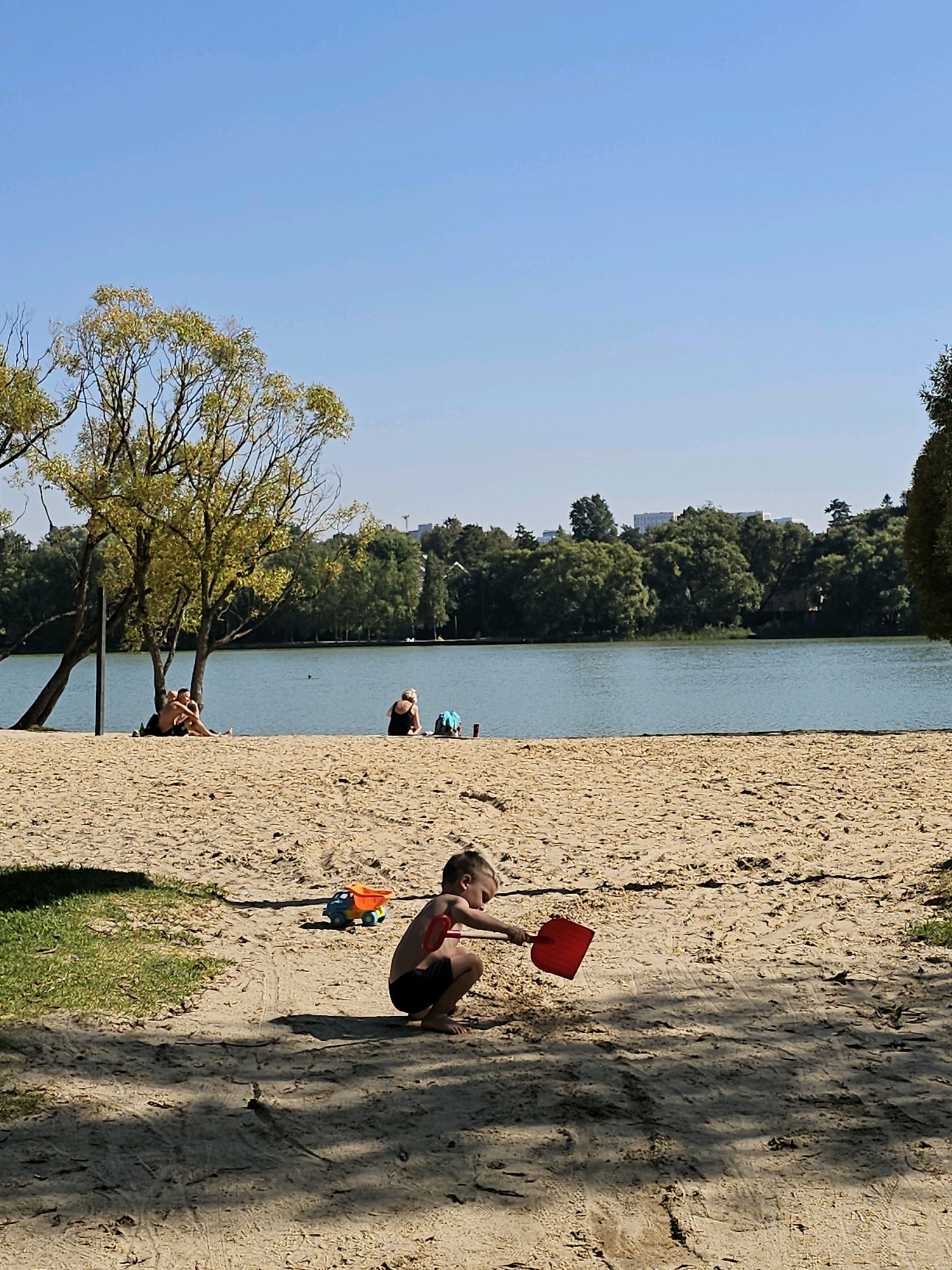 The image depicts a sunny day at a sandy beach adjacent to a calm lake. In the foreground, a young child wearing dark swim trunks is crouched down, playing with a red shovel and a colorful toy truck. The sand is marked with tire tracks and footprints. Further back on the beach, several adults are visible relaxing. Two individuals, a man and a woman, are sitting close together under the shade of a large tree. Another woman is seated with a blue towel beside her. The lake is a tranquil blue, with a few indistinct shapes in the water, possibly swimmers or boats. The background is filled with lush green trees and buildings, suggesting an urban park or recreational area. The sky is a clear, bright blue. This scene is likely at Lake Zolotoy Kolodets in Moscow, Russia, a popular spot for outdoor recreation.