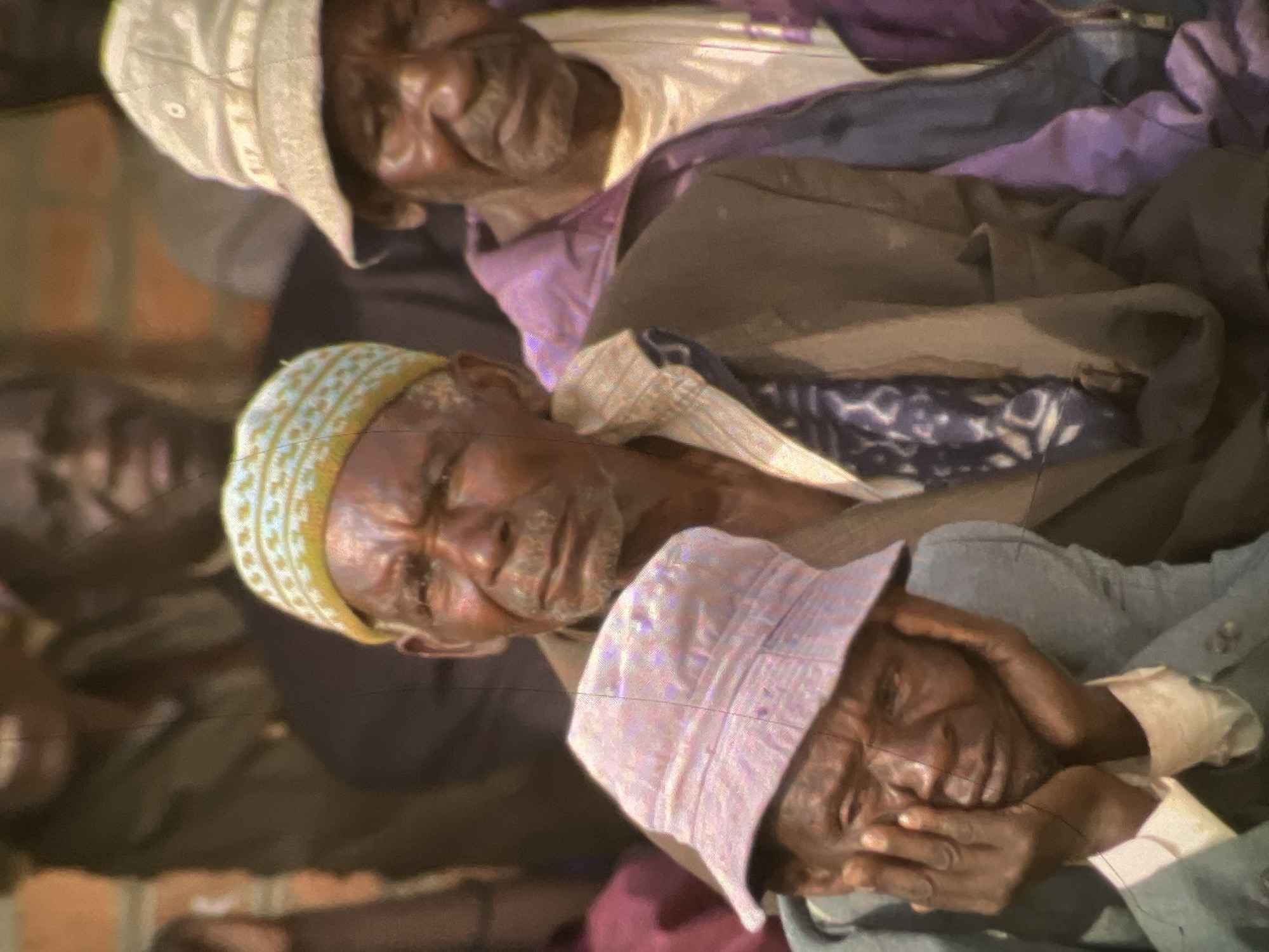 The image shows a group of people, primarily elderly men, gathered outdoors. The scene is likely a public space in Lagos, Nigeria, suggested by the presence of individuals in traditional attire and the warm, possibly humid, atmosphere. The focus is on three men in the foreground.
The man on the left, wearing a pink bucket hat and a light-colored shirt, rests his chin on his hands, his expression appearing somber or contemplative. To his right, an older man sports a light blue and yellow patterned cap, his gaze directed forward with a serious demeanor. Behind him and slightly to the right, another man wears a light-colored hat and a purple jacket over a white shirt, looking towards the side. The background shows other individuals, some out of focus, suggesting a gathering or a community event. The lighting is soft, and the overall tone of the image is muted, possibly due to the photographic technique or the natural light conditions.