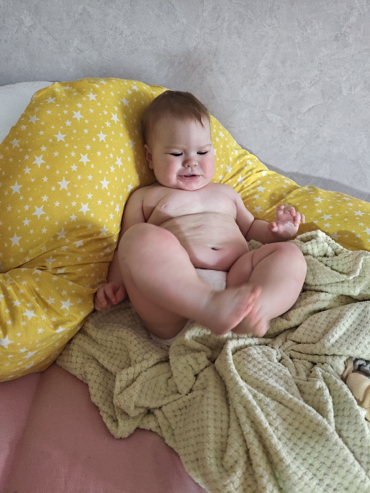 The image shows an infant seated indoors in Chaikovsky, Russia. The light-skinned baby, with light brown hair, is bare-chested and supported by a yellow pillow adorned with white star patterns. A light green, textured blanket covers the infant's legs, which are bent, with the feet blurred from motion. A pink surface is visible beneath the blanket. The background consists of a textured light gray wall. The infant displays a content facial expression with narrowed eyes.