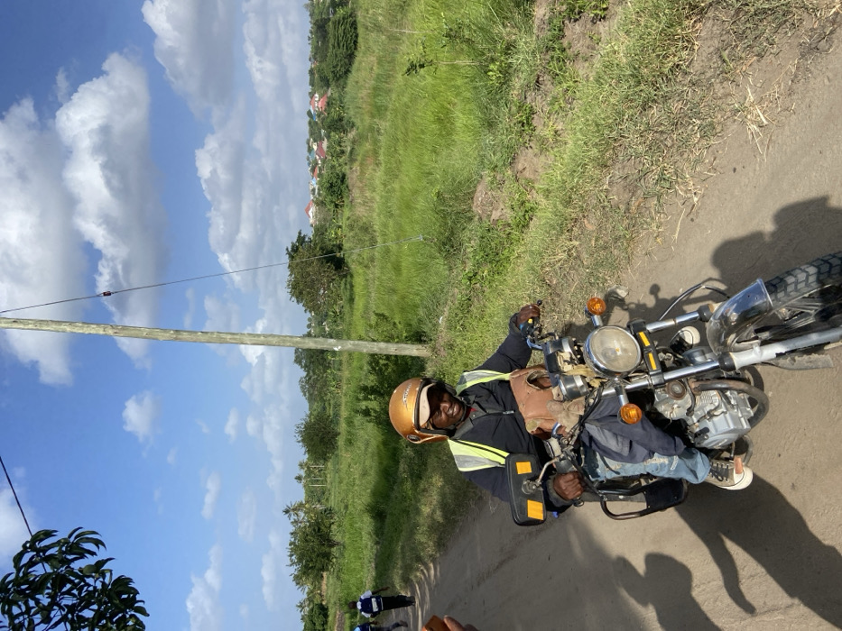A man wearing a helmet rides a motorcycle on a dirt road. He is wearing a safety vest and light-colored sneakers. A power pole stands to his left, with a field of grass and some houses in the background. The sky is blue with scattered clouds. Another person can be seen partially in the lower left-hand corner.